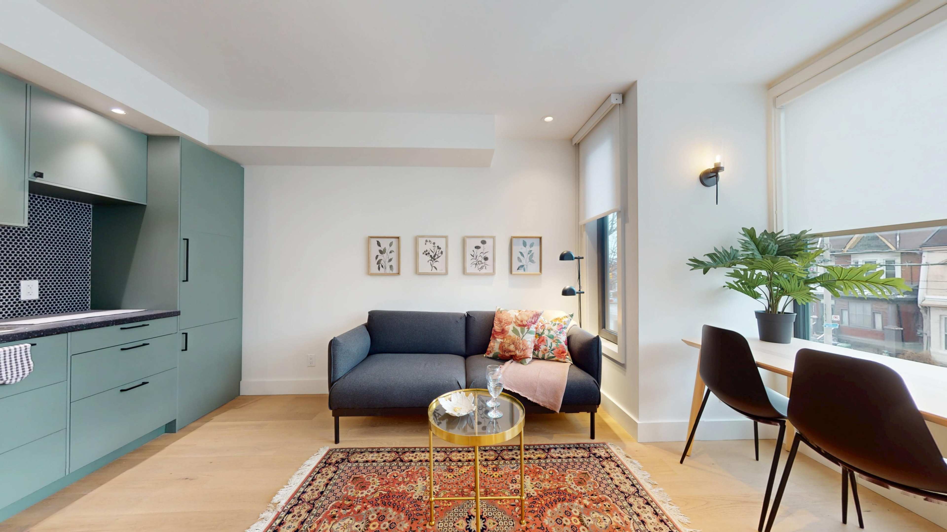 The image shows a modern, well-lit living area featuring a blue sofa, a round gold coffee table, and a small dining set next to a large window.