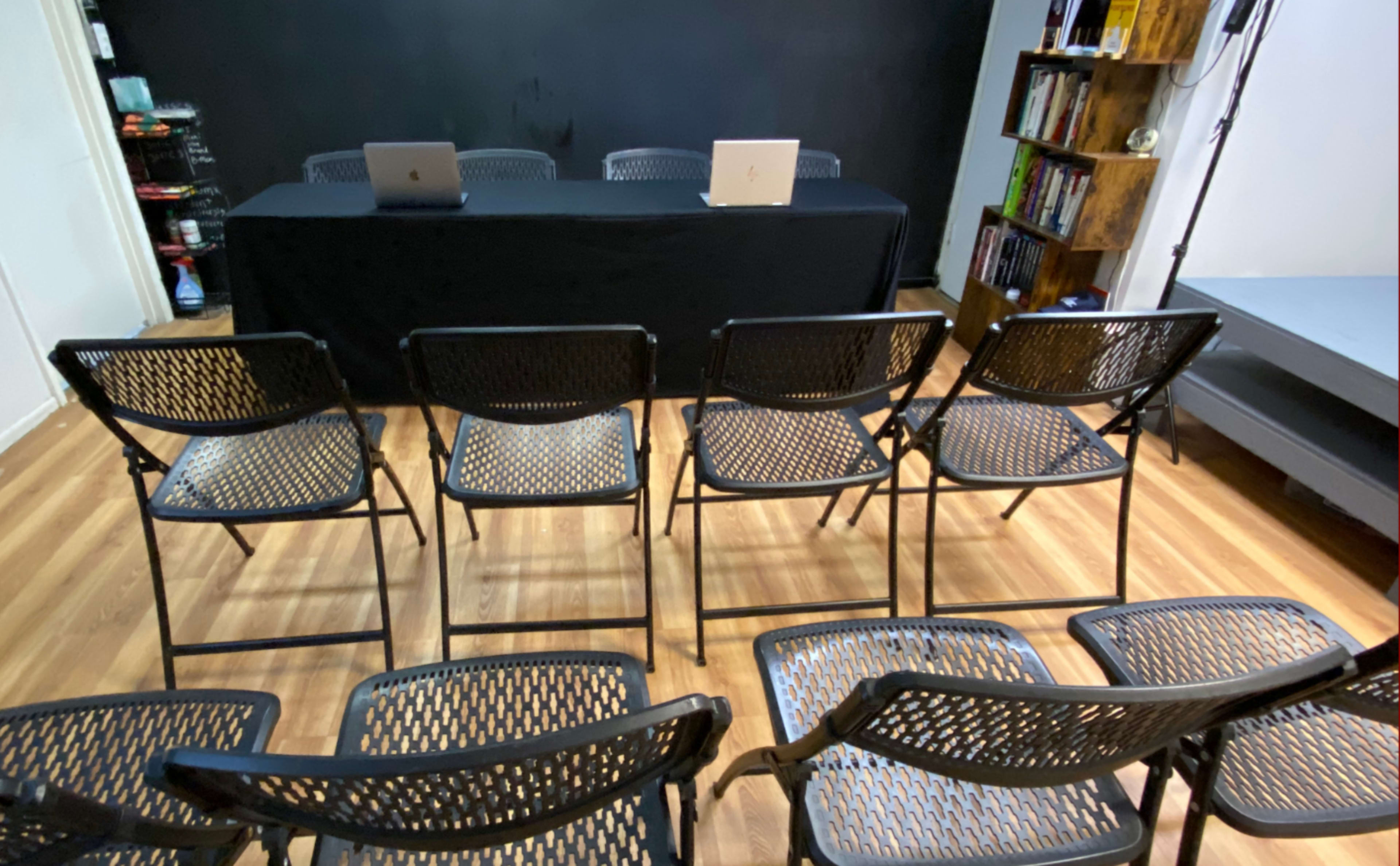 A row of black folding chairs faces a table with two laptops against a dark wall.