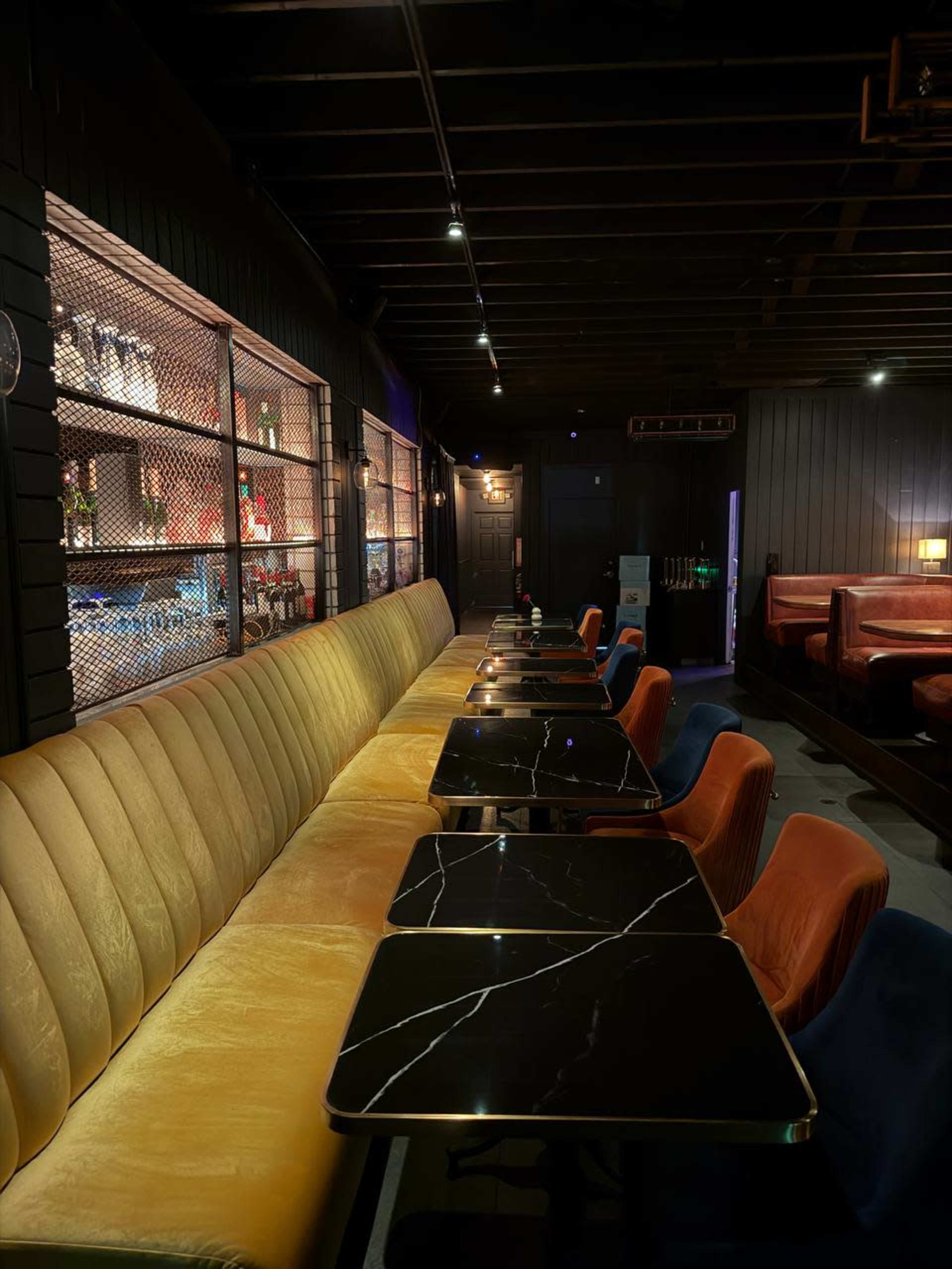 The image shows a dimly lit lounge area with a long, upholstered yellow bench and several tables featuring dark, marble-like surfaces.