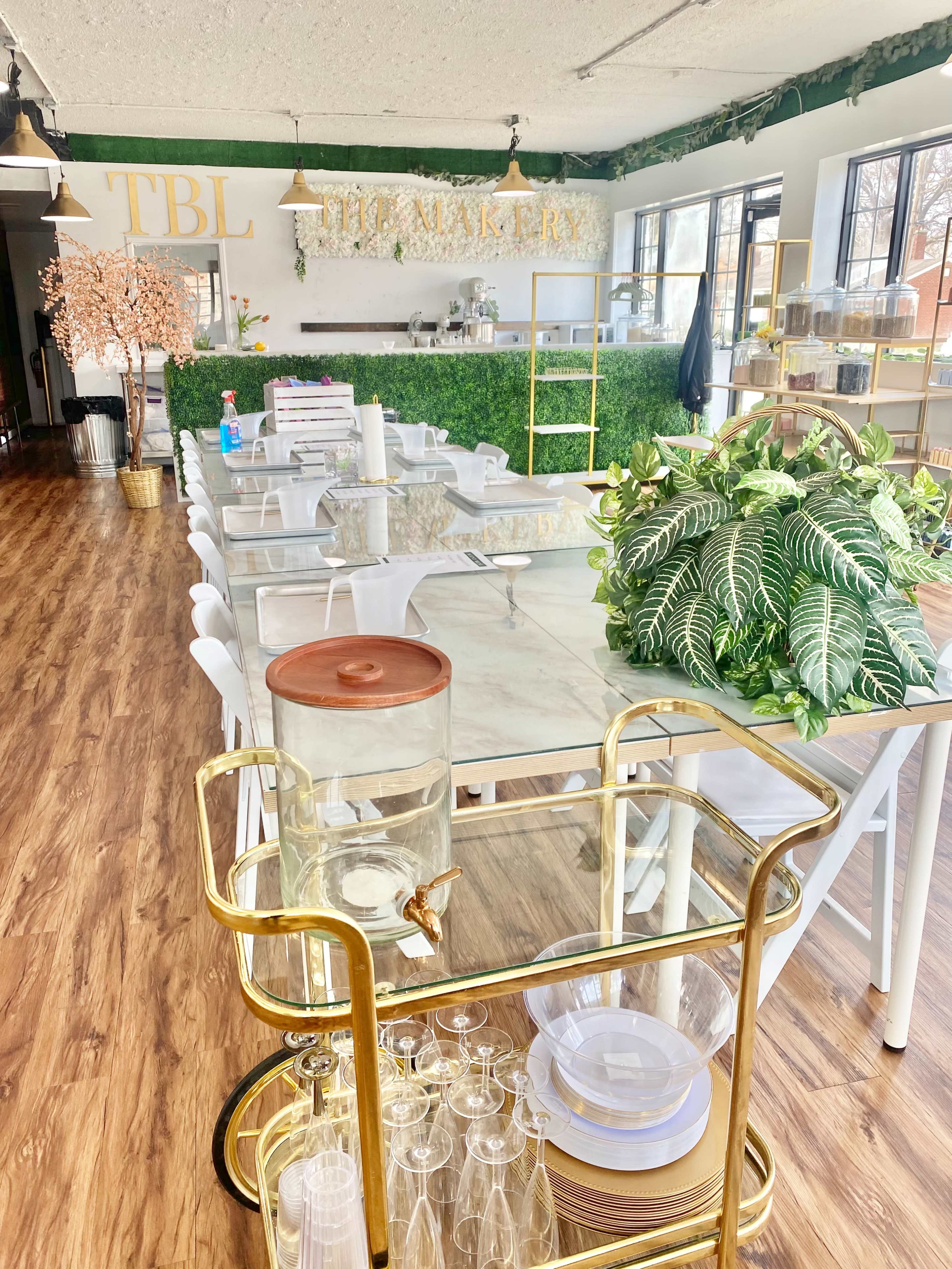 A spacious, elegantly designed café or bakery interior with a long table, greenery on the walls, and a cart displaying glassware and refreshments.