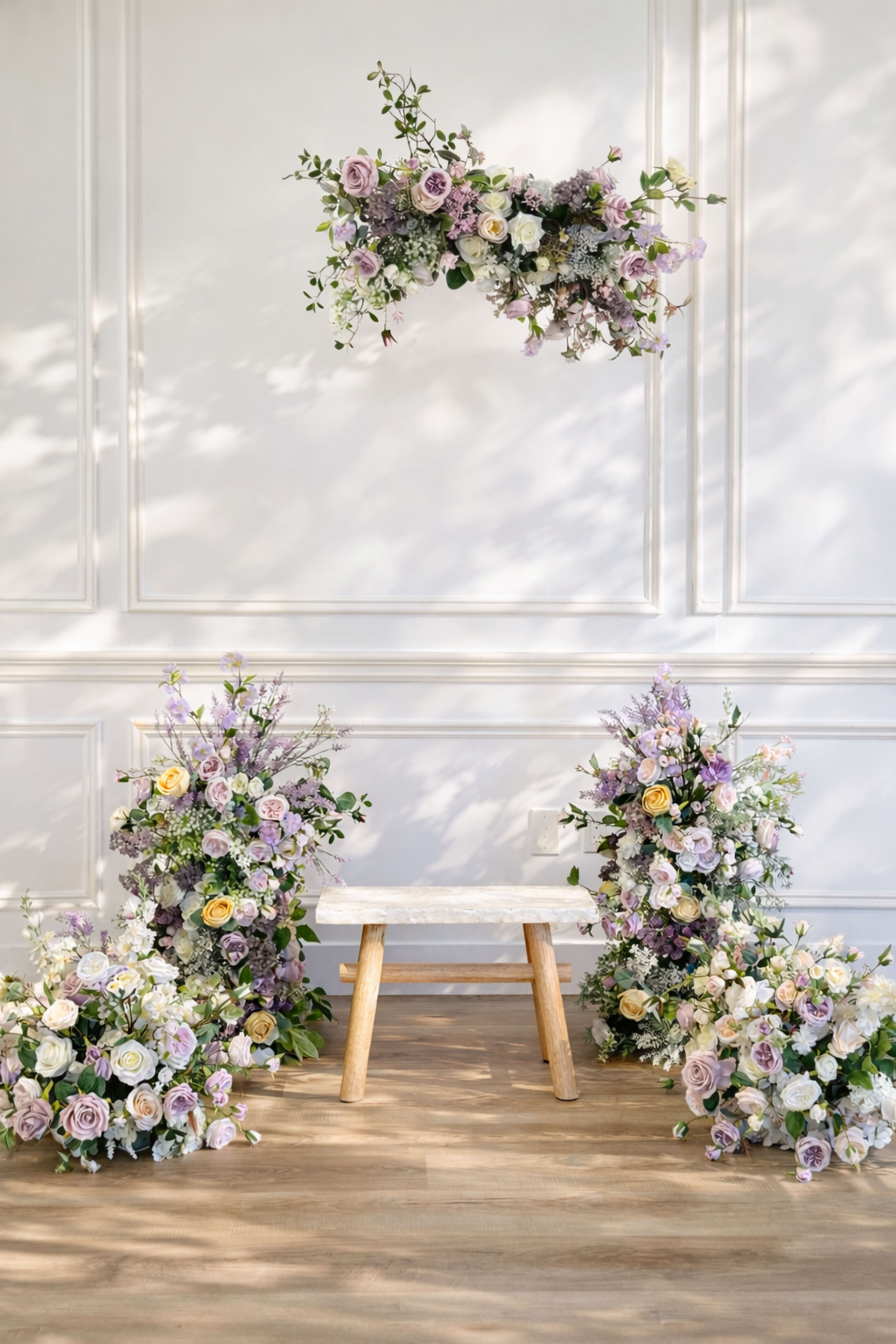 The scene features a simple wooden stool surrounded by large floral arrangements in shades of purple, yellow, and white, against a white wall with molded panels.