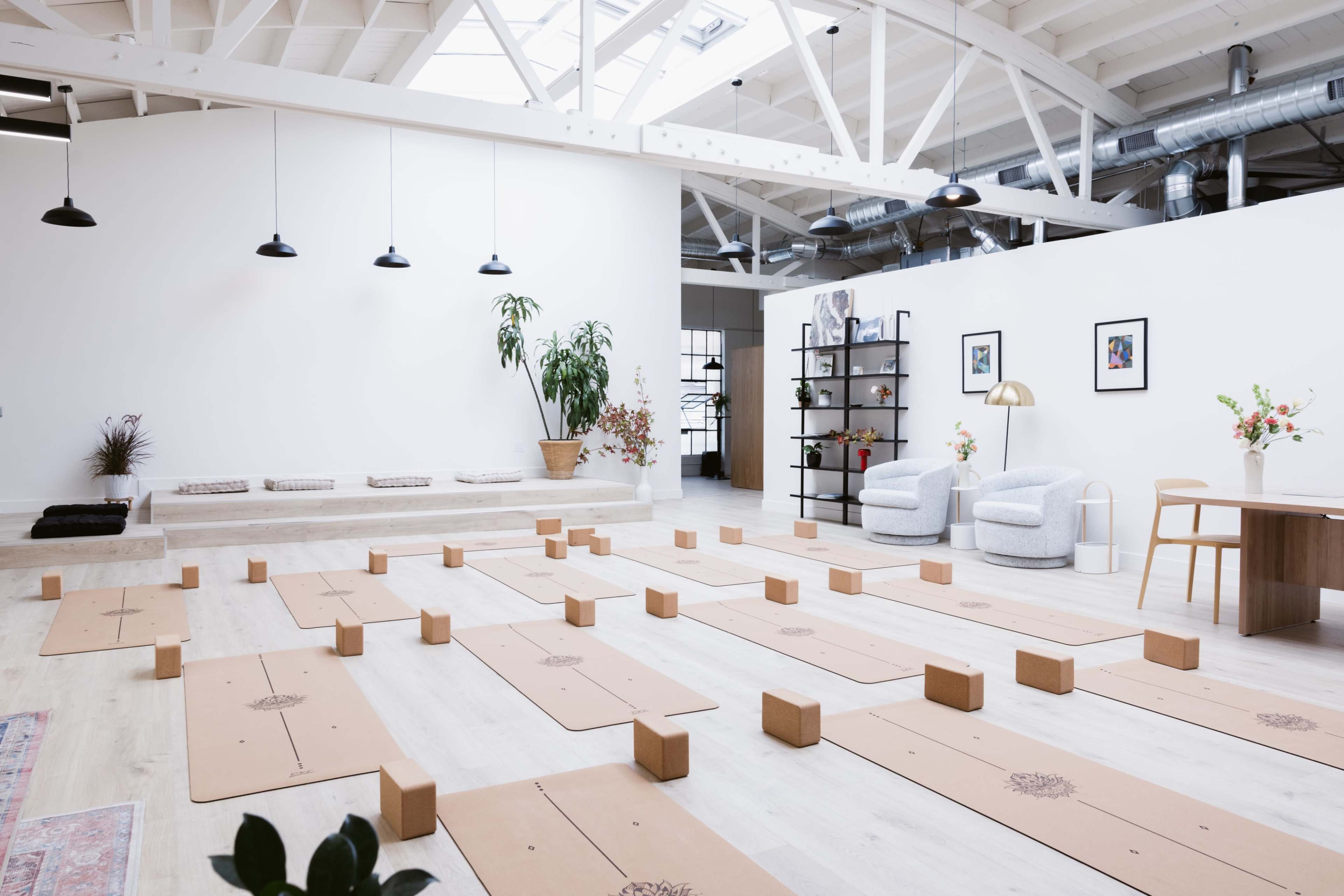 The image shows a bright, open yoga studio with neatly arranged yoga mats and wooden blocks, along with a seating area and decorative plants.