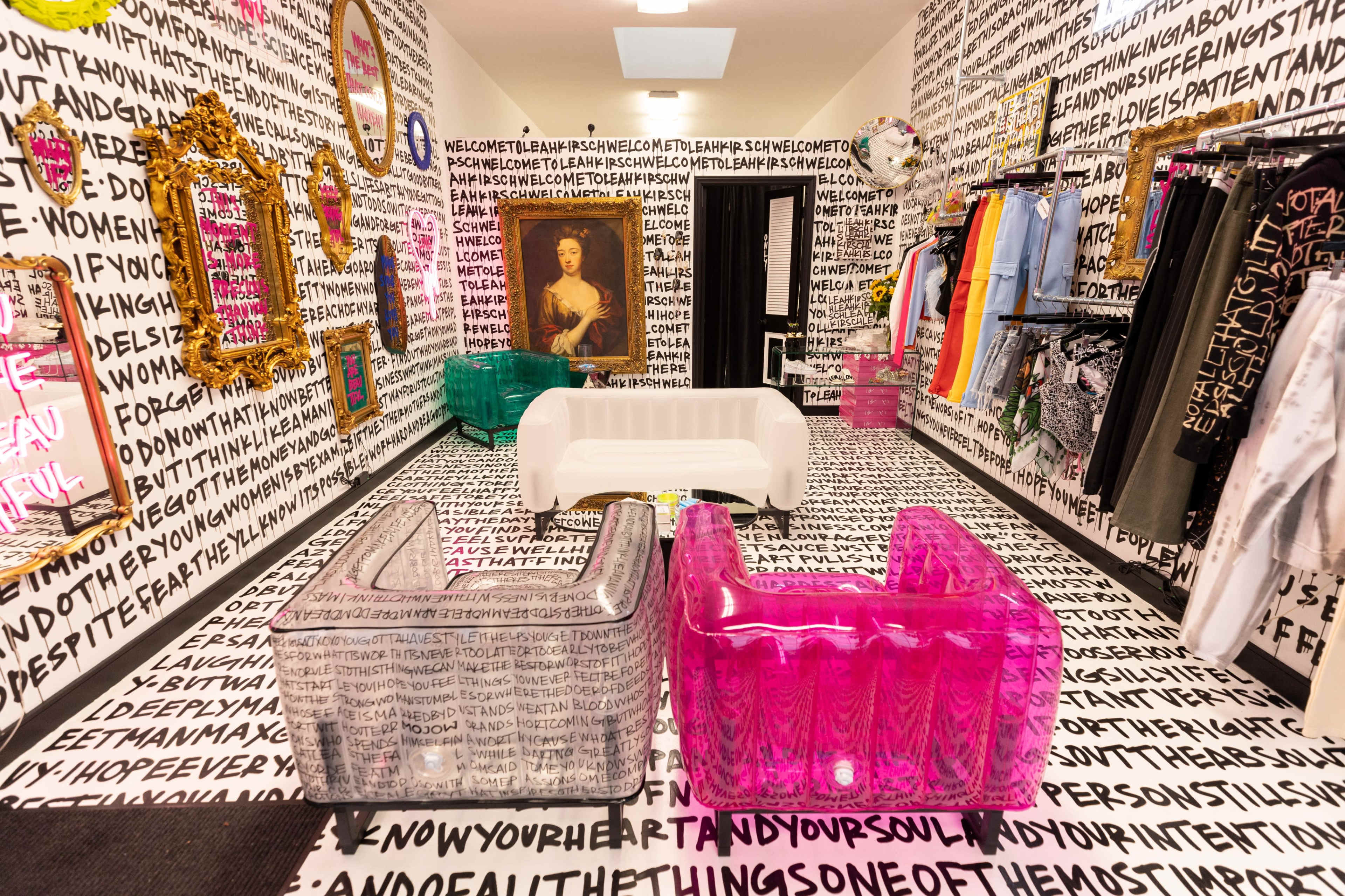A vibrant boutique interior with walls and flooring covered in repetitive text, featuring colorful transparent furniture and various clothing items displayed on racks.