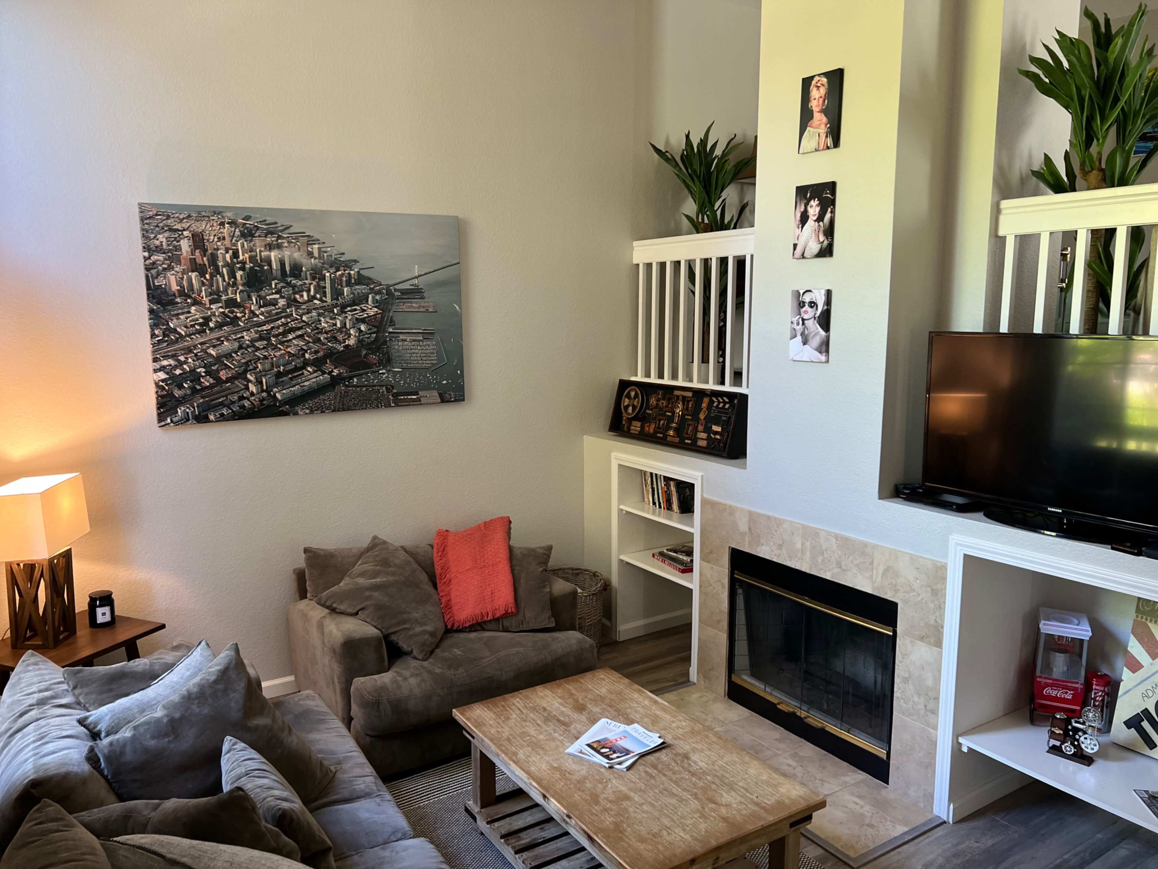 A cozy living room with a large cityscape painting, a comfortable sofa arrangement, a wooden coffee table, and a television set next to a fireplace.