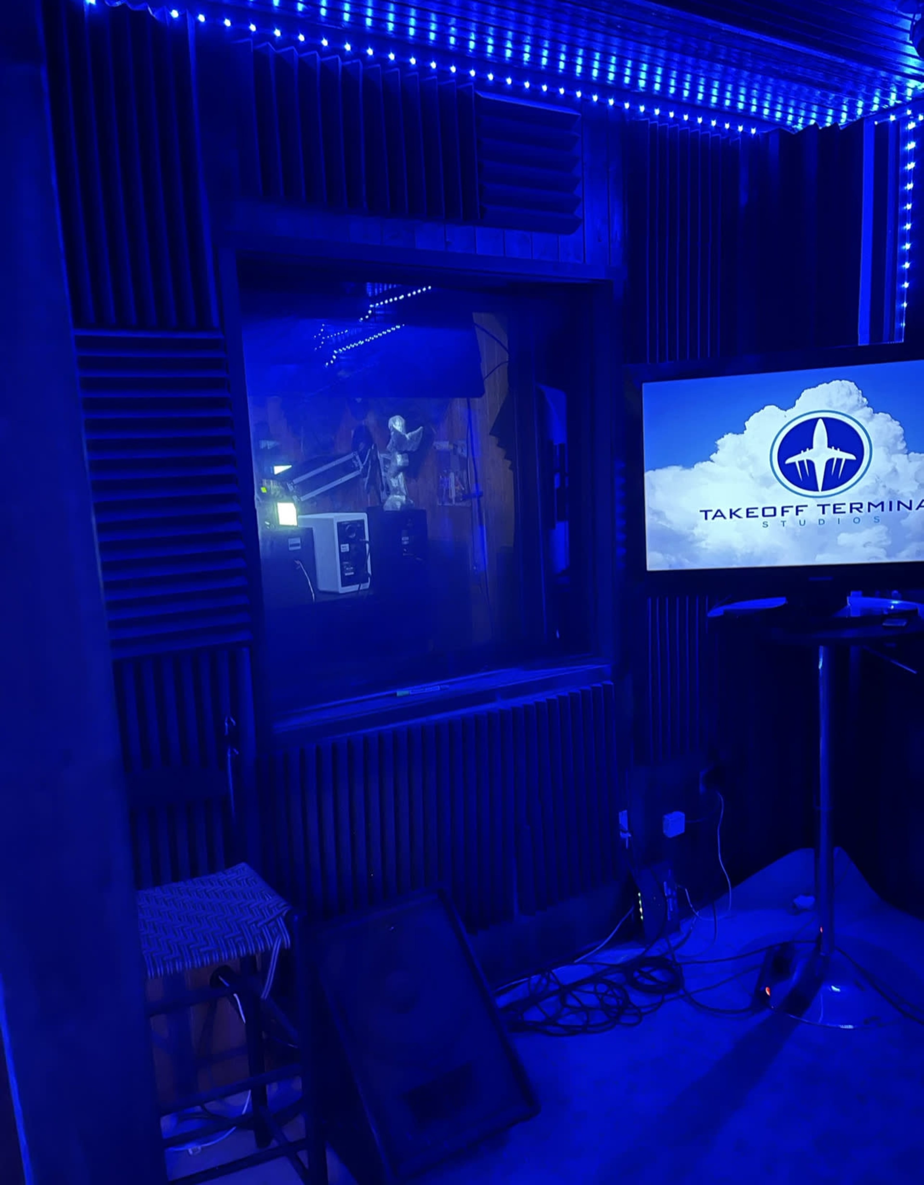 A recording studio with blue lighting, featuring a large window, a television displaying "TAKEOFF TERMINI," and a stool next to a sound system.