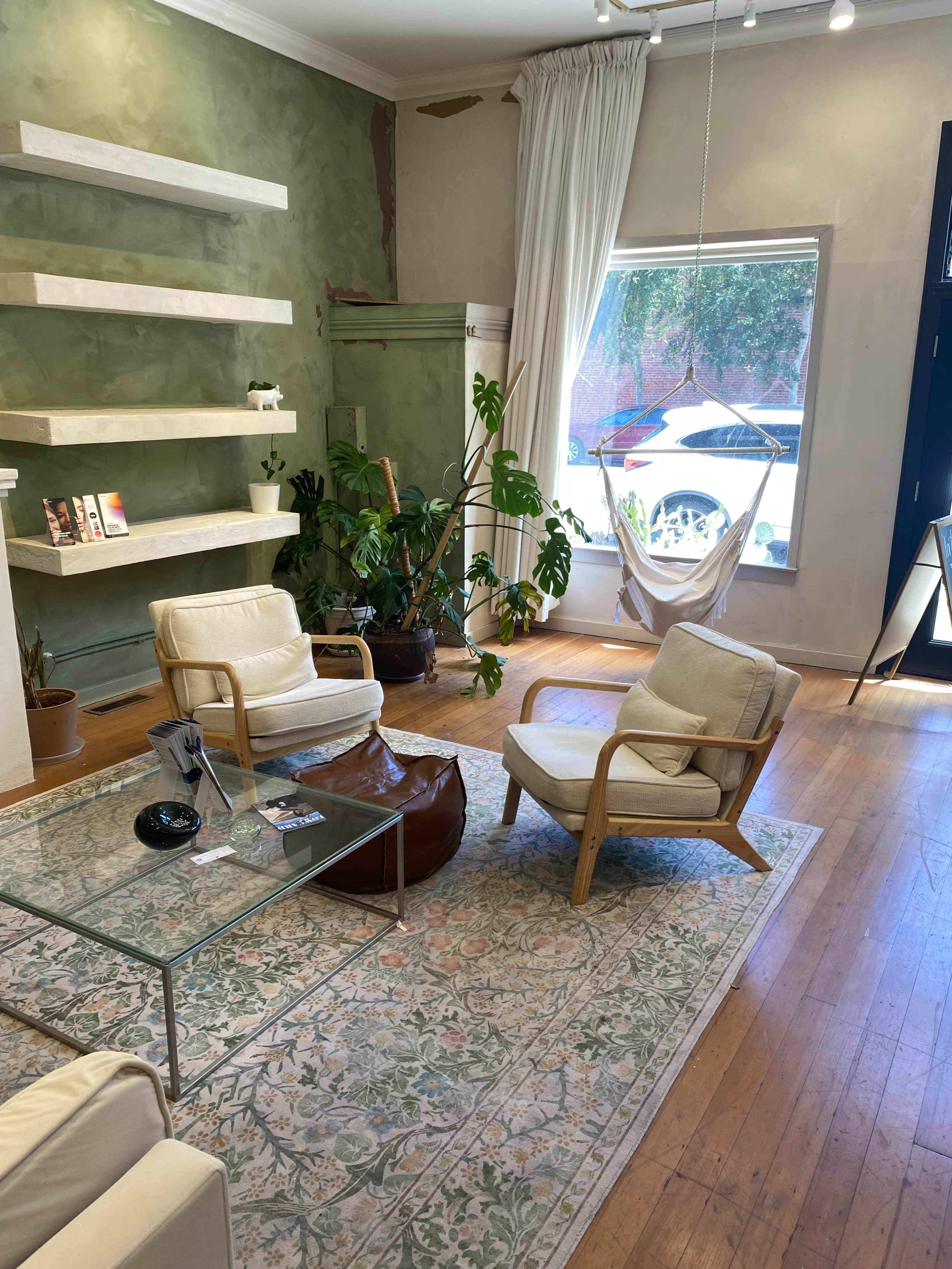 The image shows a cozy interior space with two white armchairs, a glass coffee table, plants, a hammock, and shelves on a green wall.