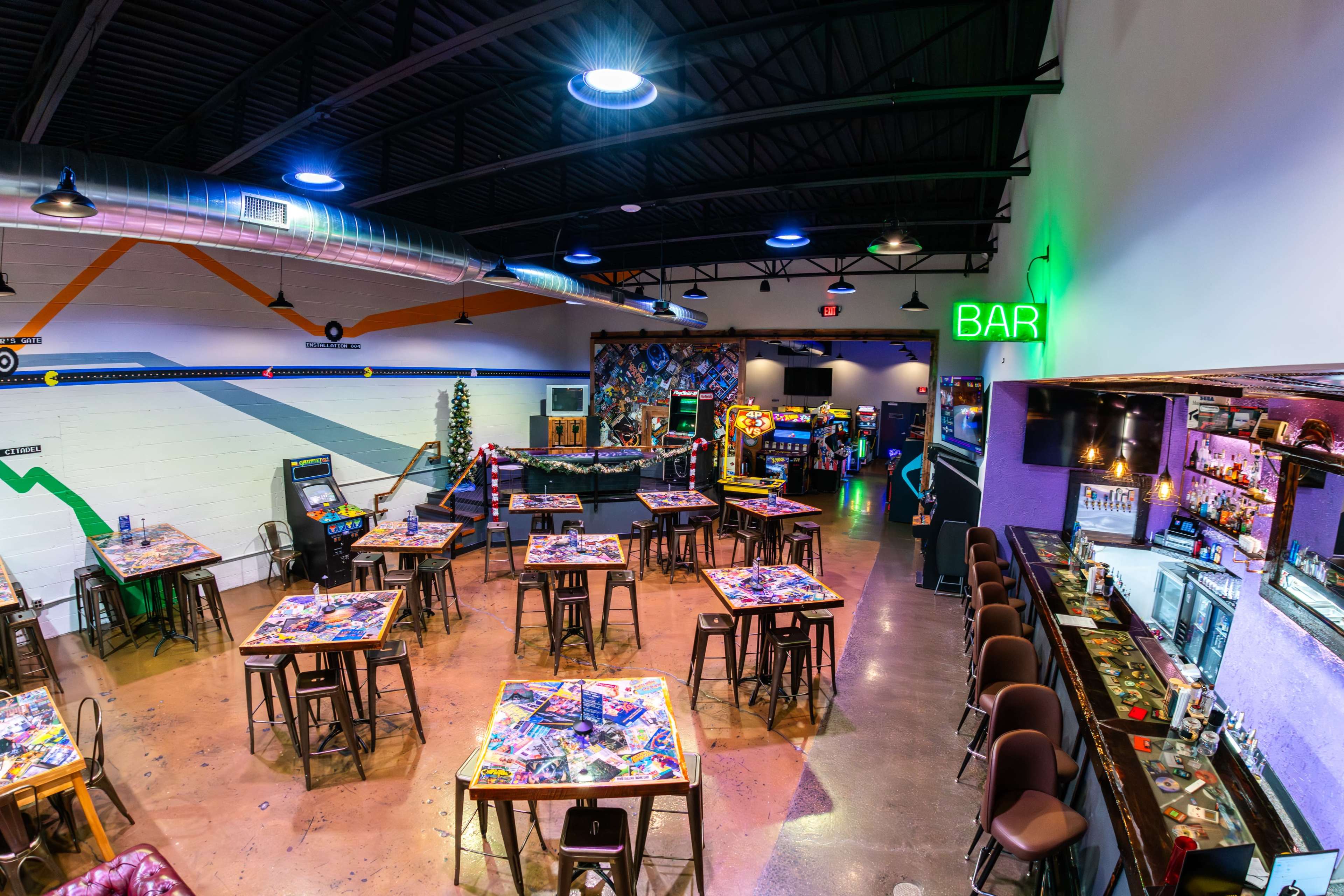 The image shows a vibrant arcade with various tables and gaming machines, alongside a bar area illuminated by green lights.