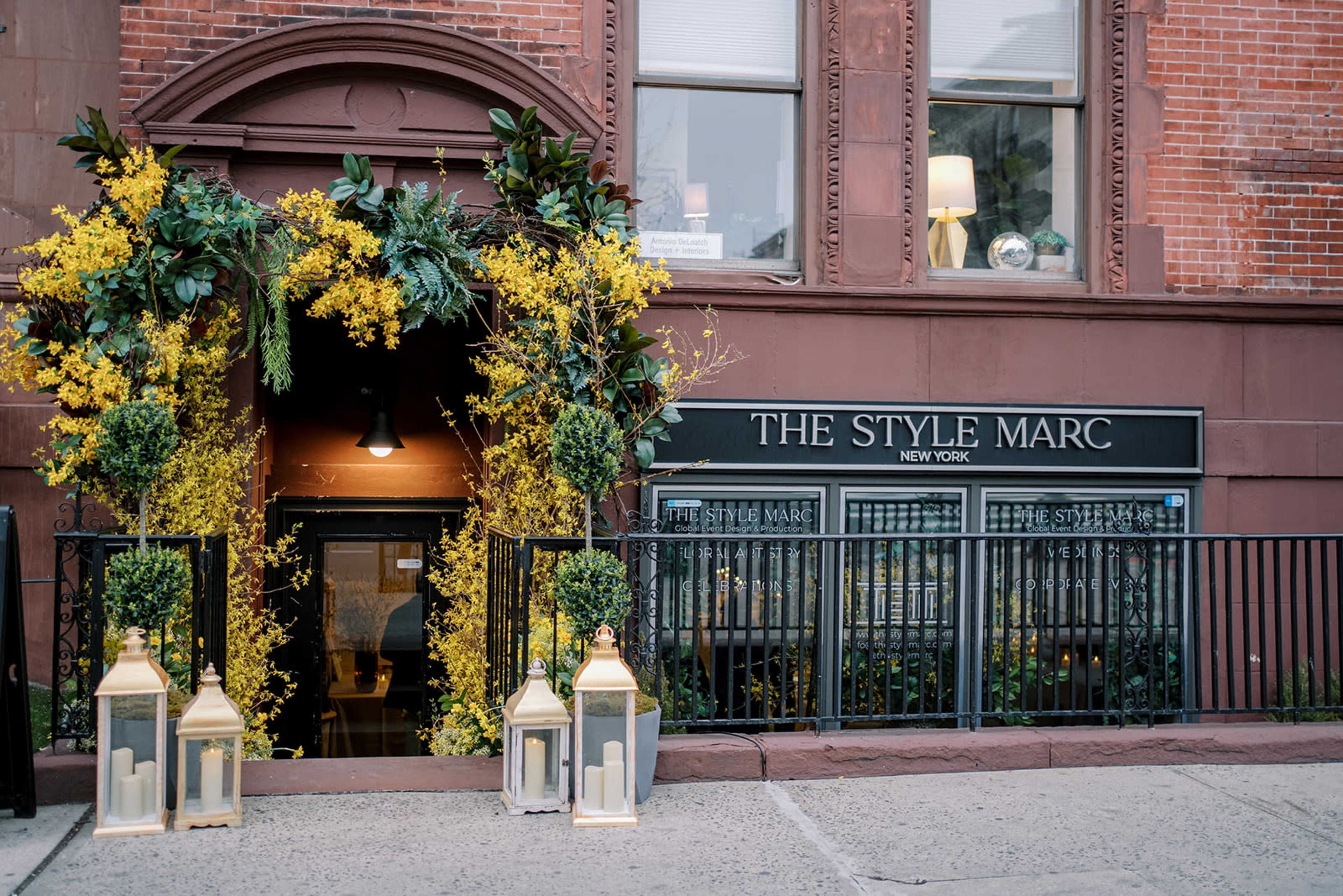 The entrance of The Style Marc features a decorative arch of yellow flowers and greenery, flanked by lanterns, against a brick facade.
