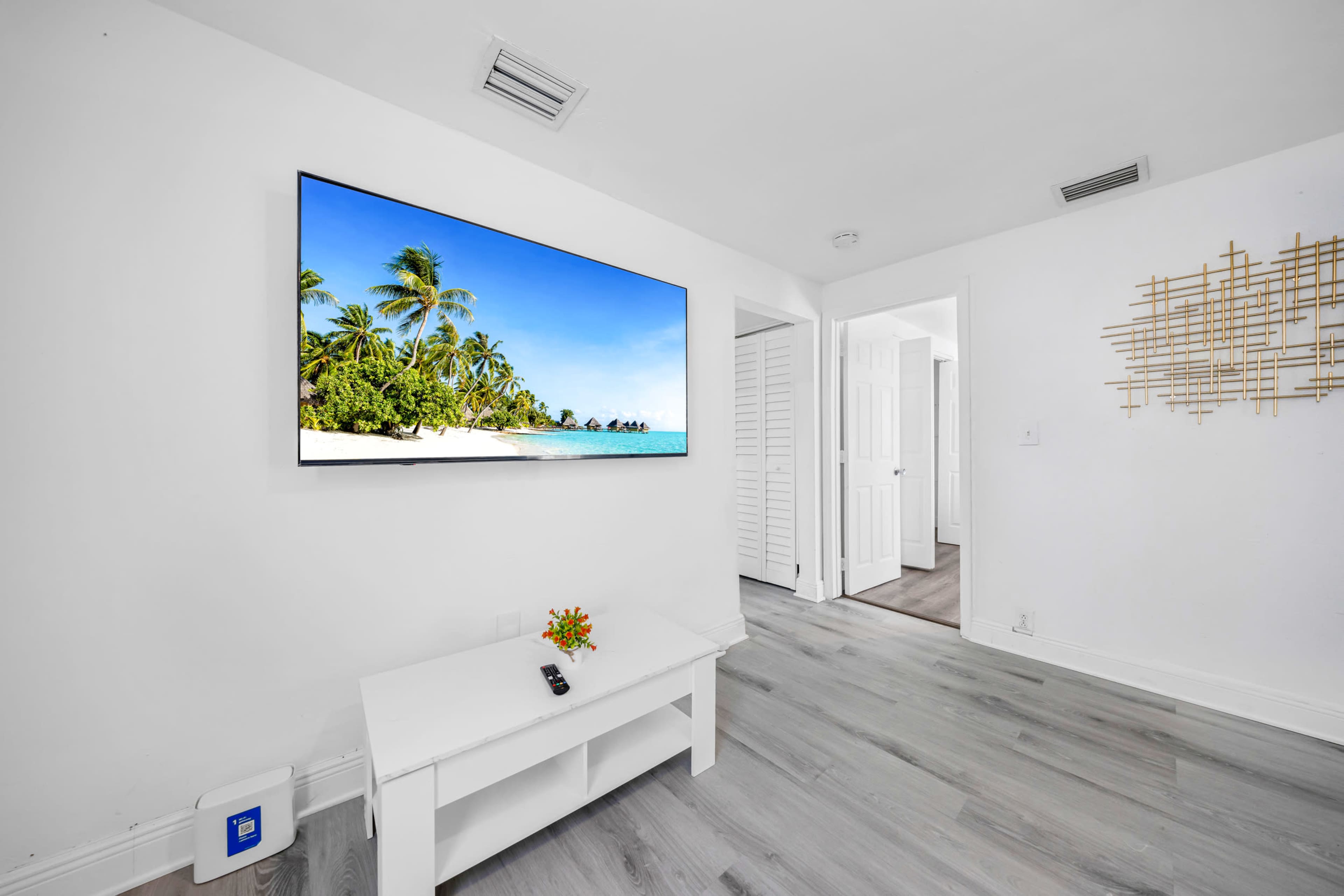 A modern living room features a large television mounted on the wall displaying a beach scene, with a white coffee table in front and an open door leading to another room.