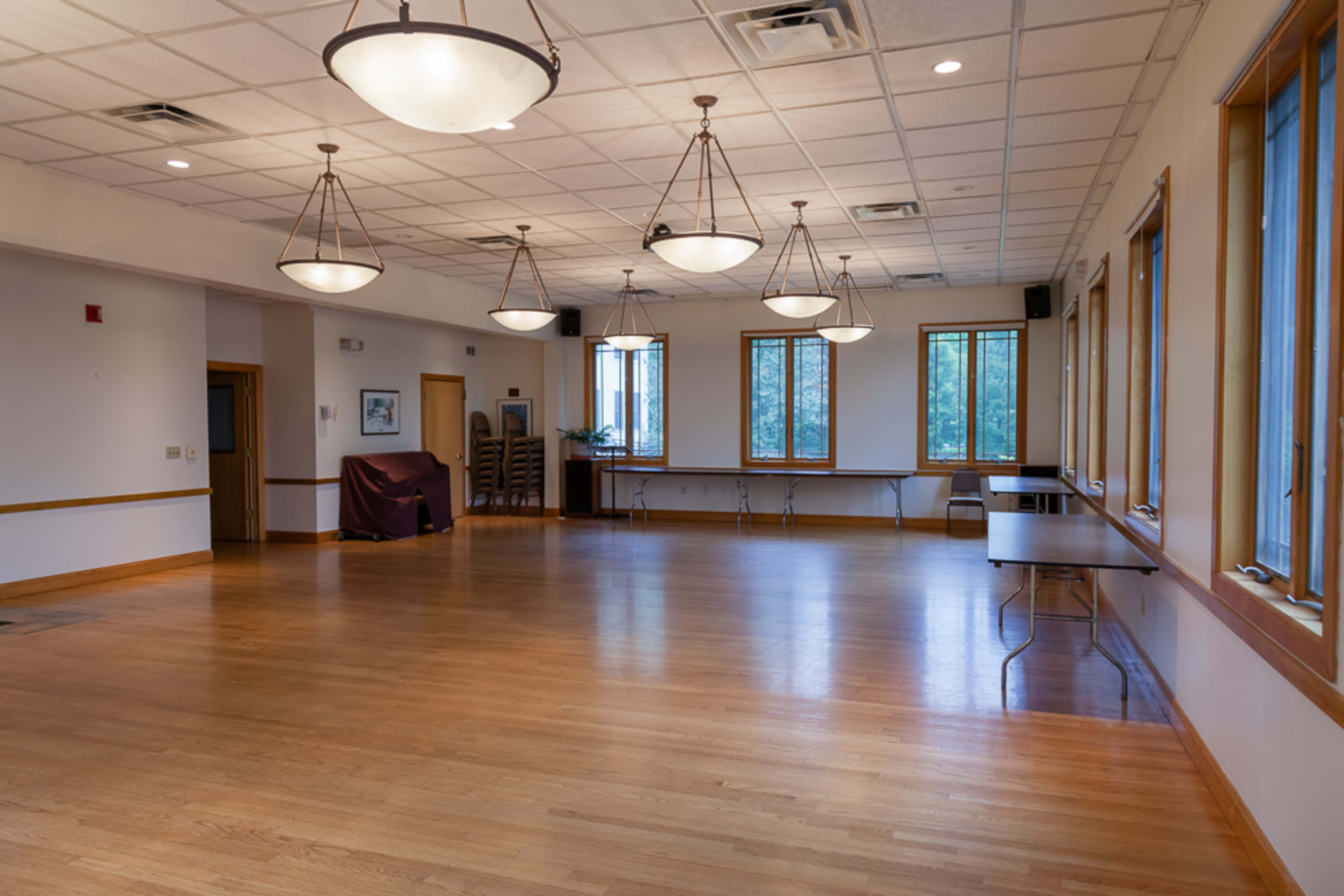 The image shows a spacious, well-lit room with wooden flooring, six hanging light fixtures, and multiple windows along the walls.