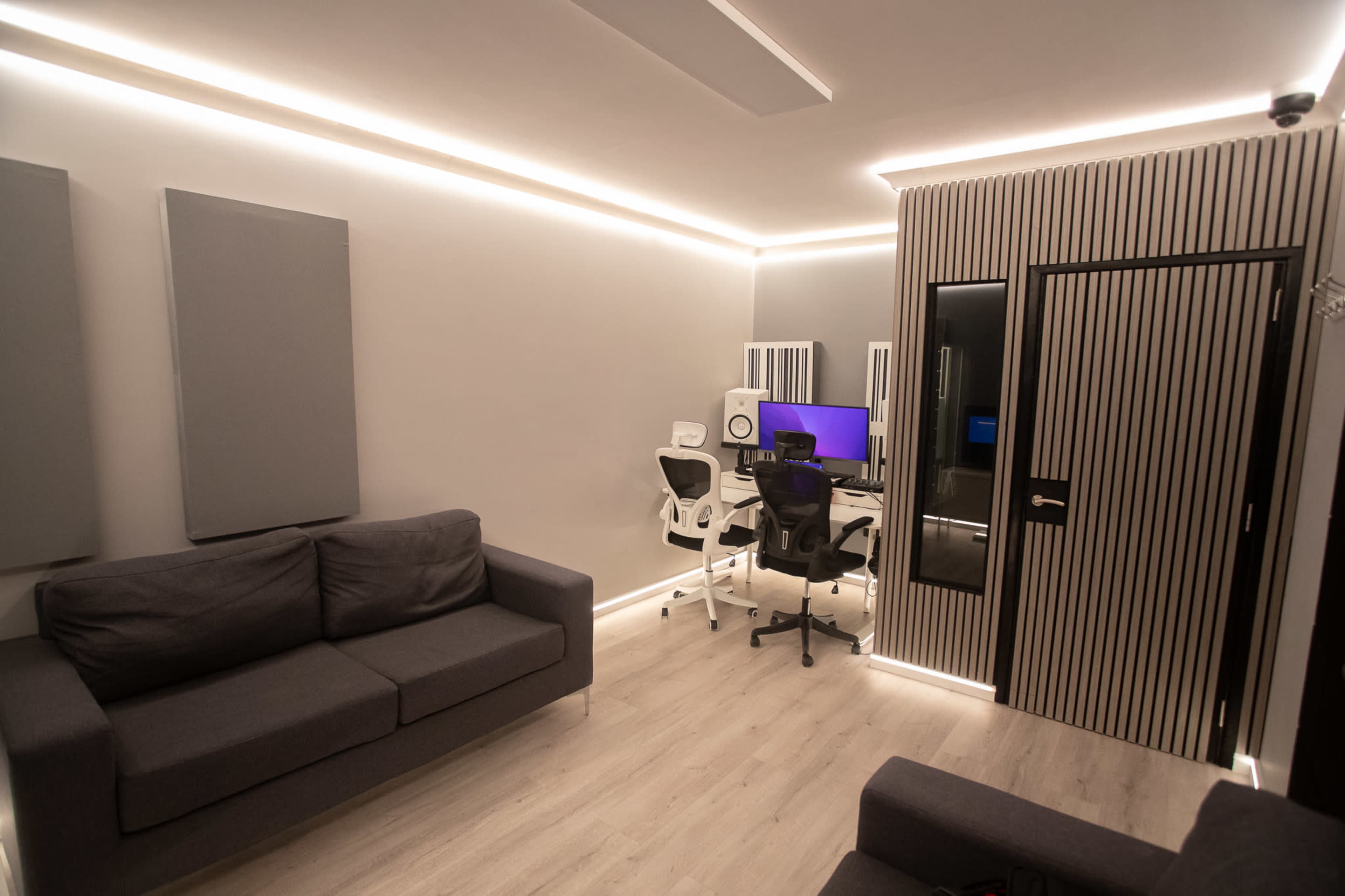 The room features a modern workspace with a desk and chair, acoustic panels on the walls, and a sofa, all illuminated by sleek lighting.