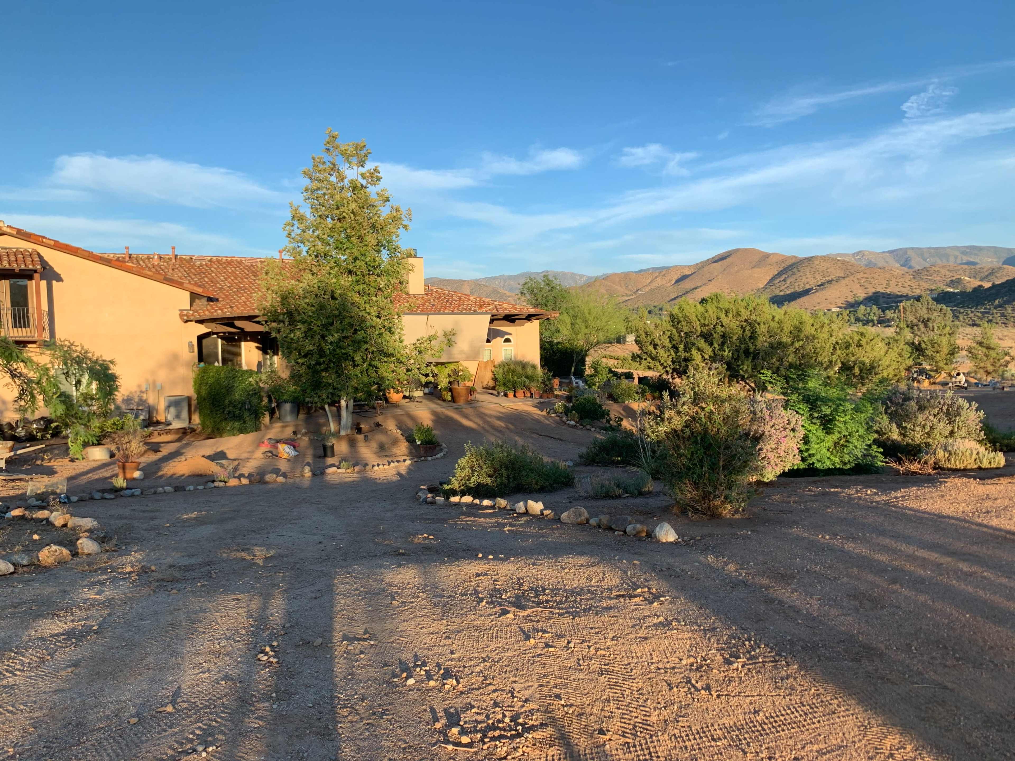 A desert house with a tiled roof and surrounded by sparse vegetation sits against a backdrop of mountains under a clear sky.