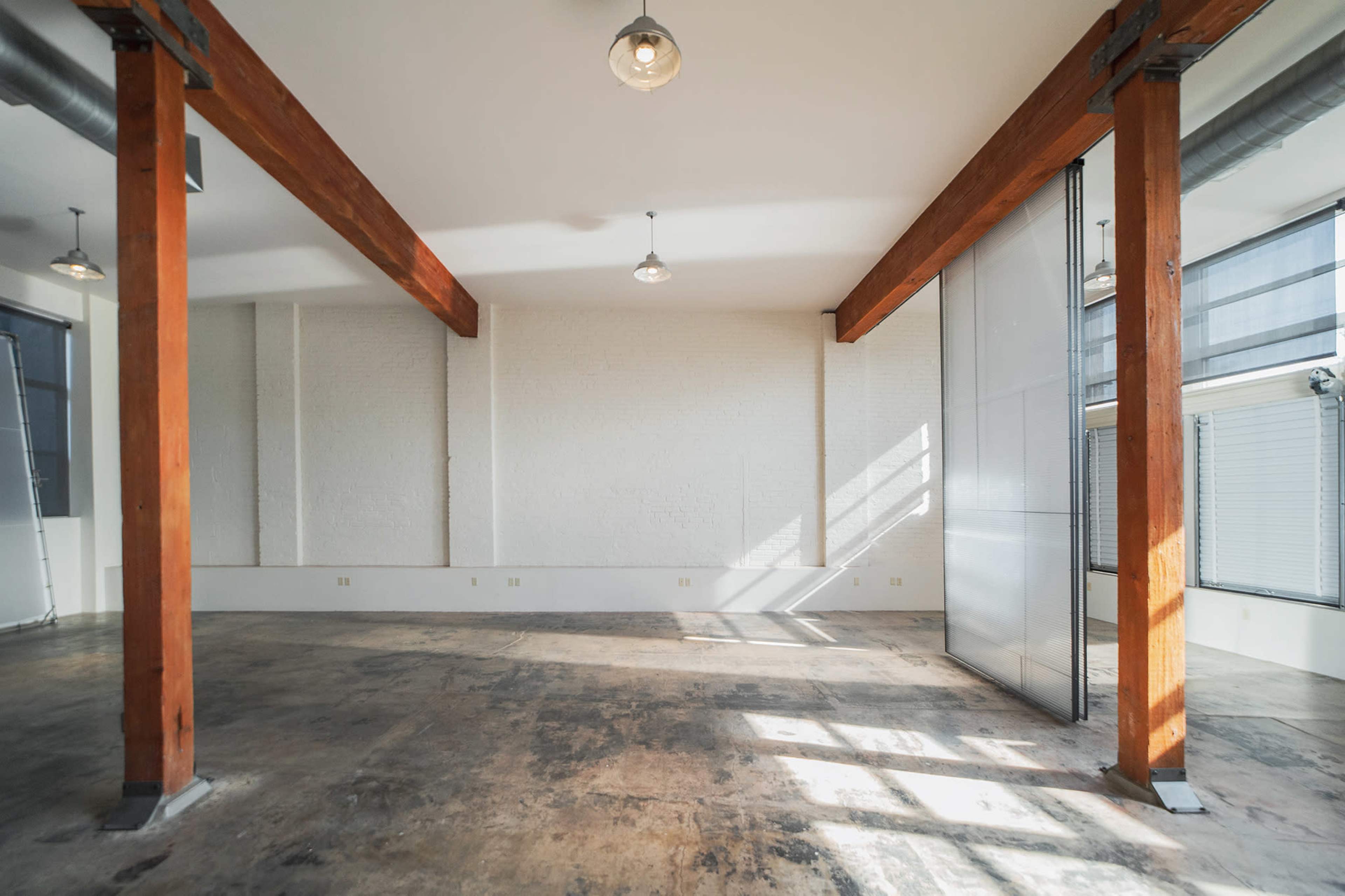 An empty room with exposed wooden beams, large windows, and a concrete floor.