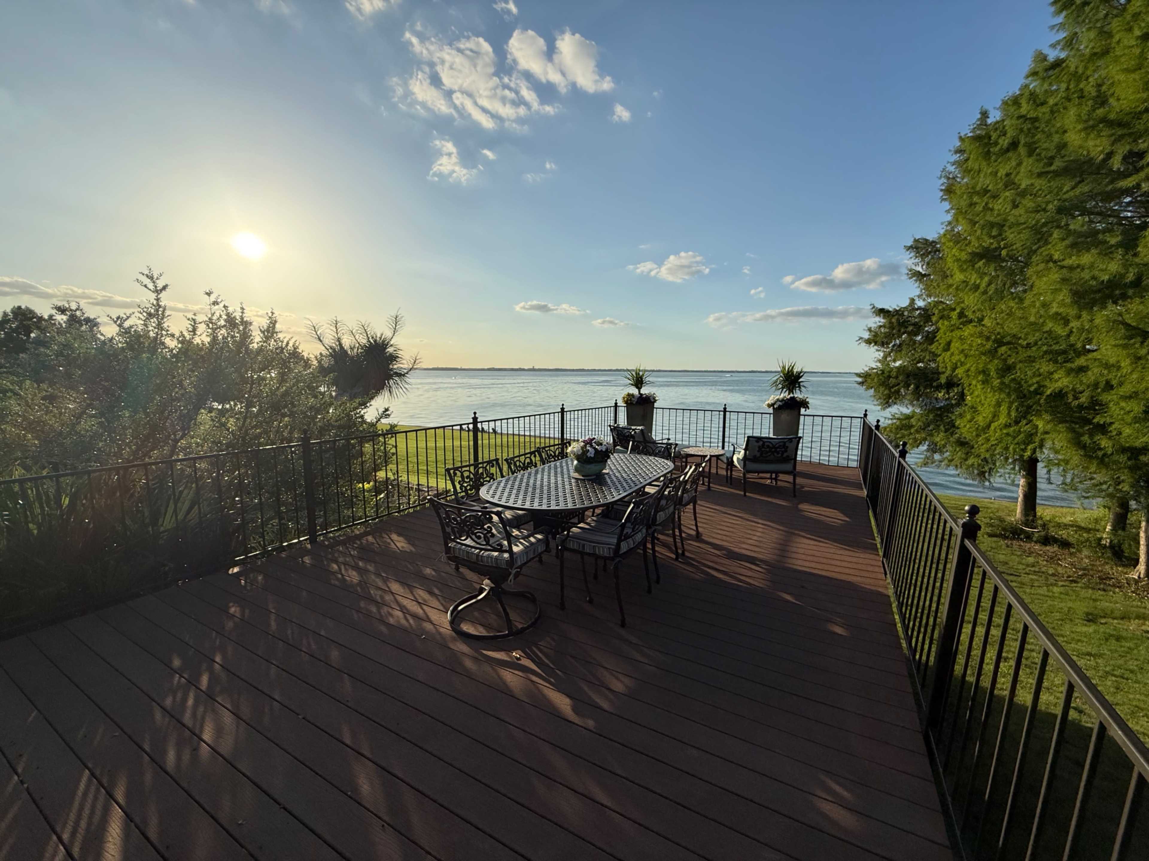 A wooden deck with a metal table and chairs overlooks a calm body of water under a blue sky.