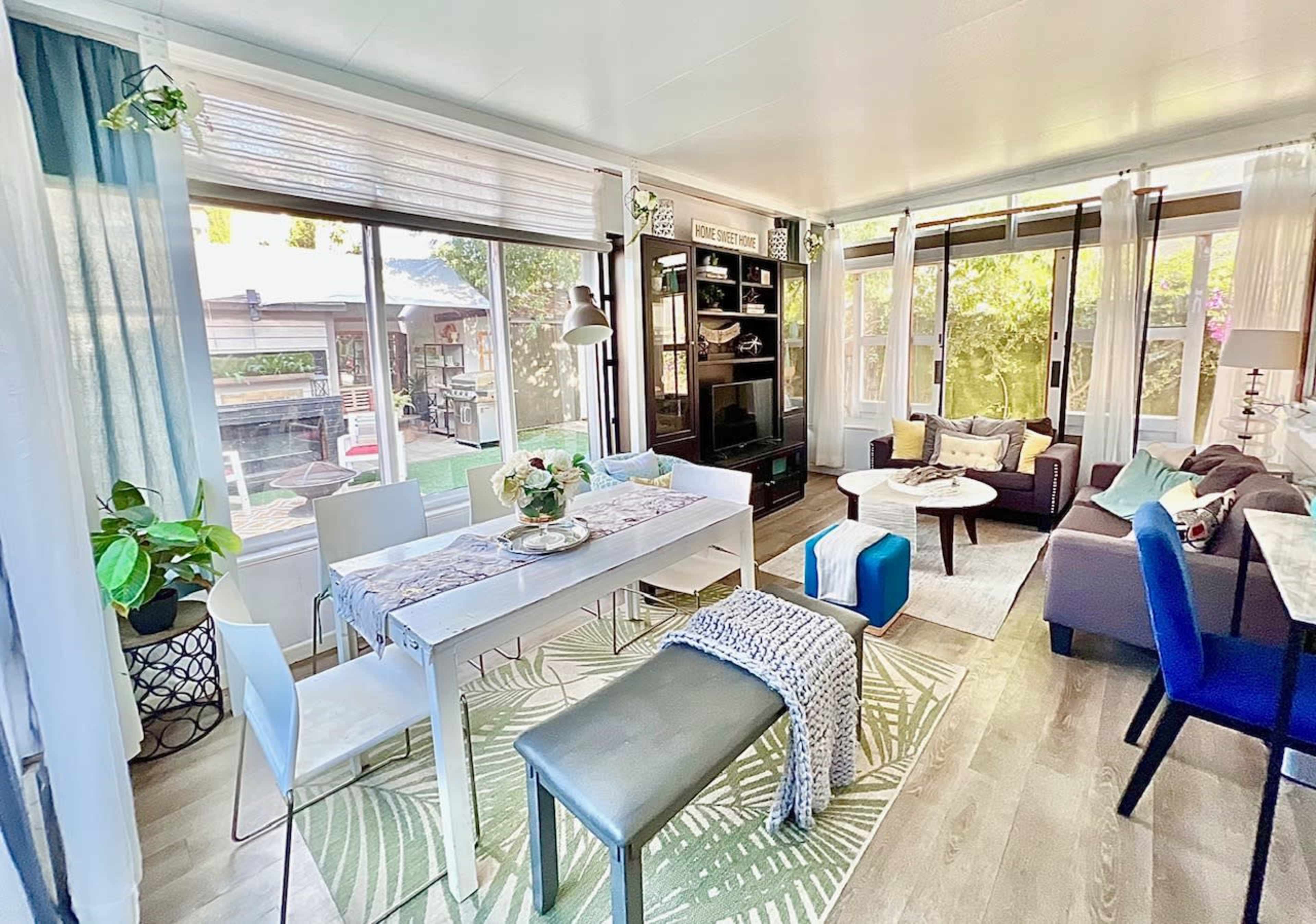 A brightly lit living area with a dining table, a sectional sofa, and large windows allowing natural light to enter.