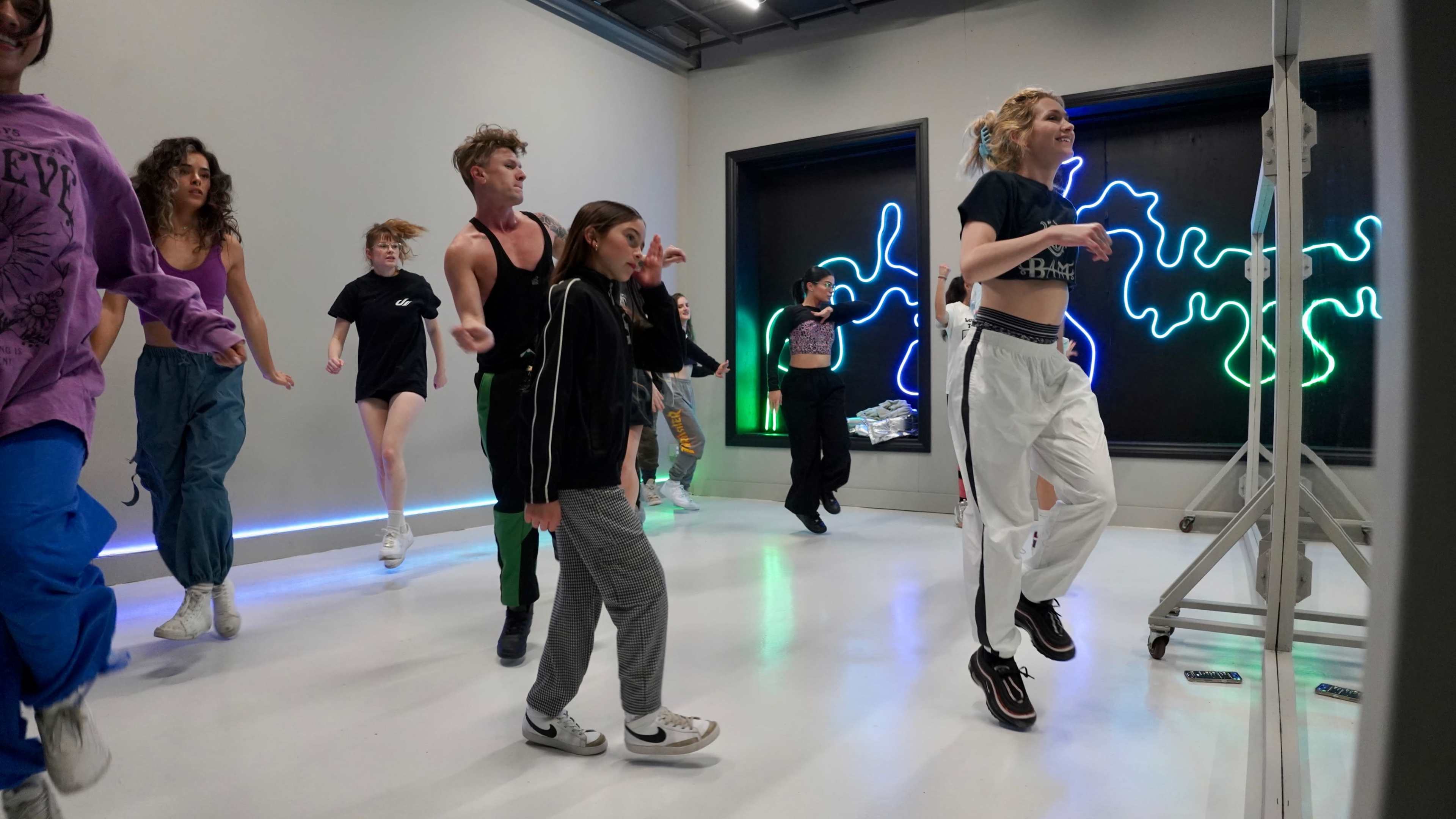 A group of dancers is practicing in a well-lit studio with neon decorations on the walls.