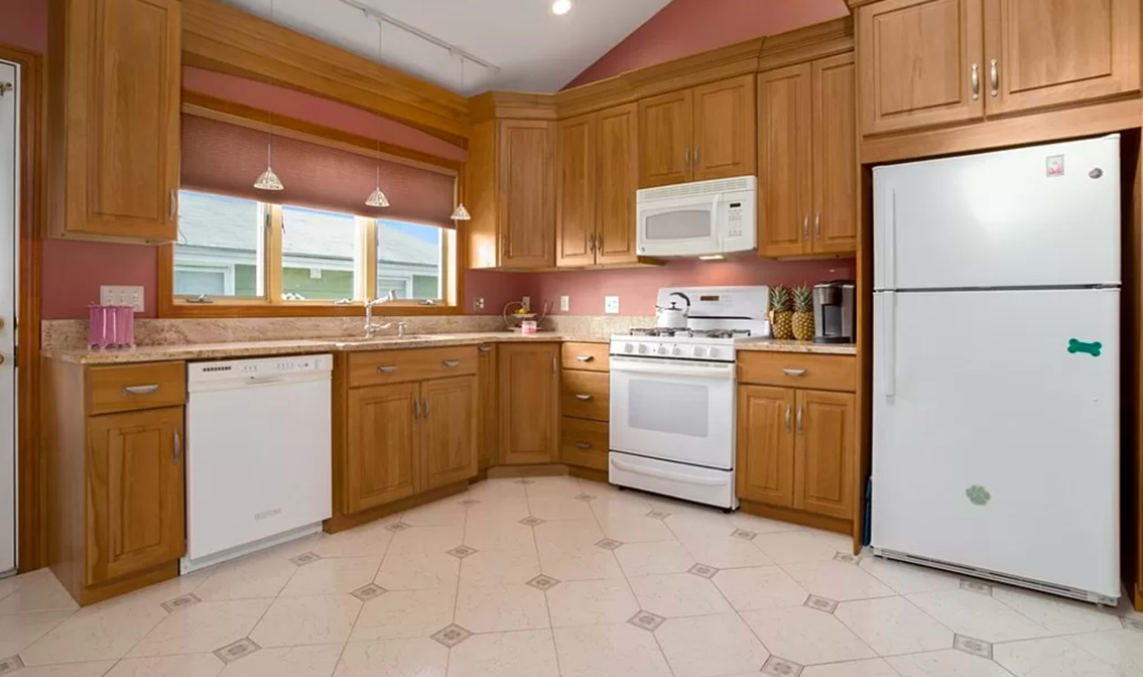 The image shows a kitchen with wooden cabinetry, a white refrigerator, a stove, and a dishwasher, set against a pink wall.