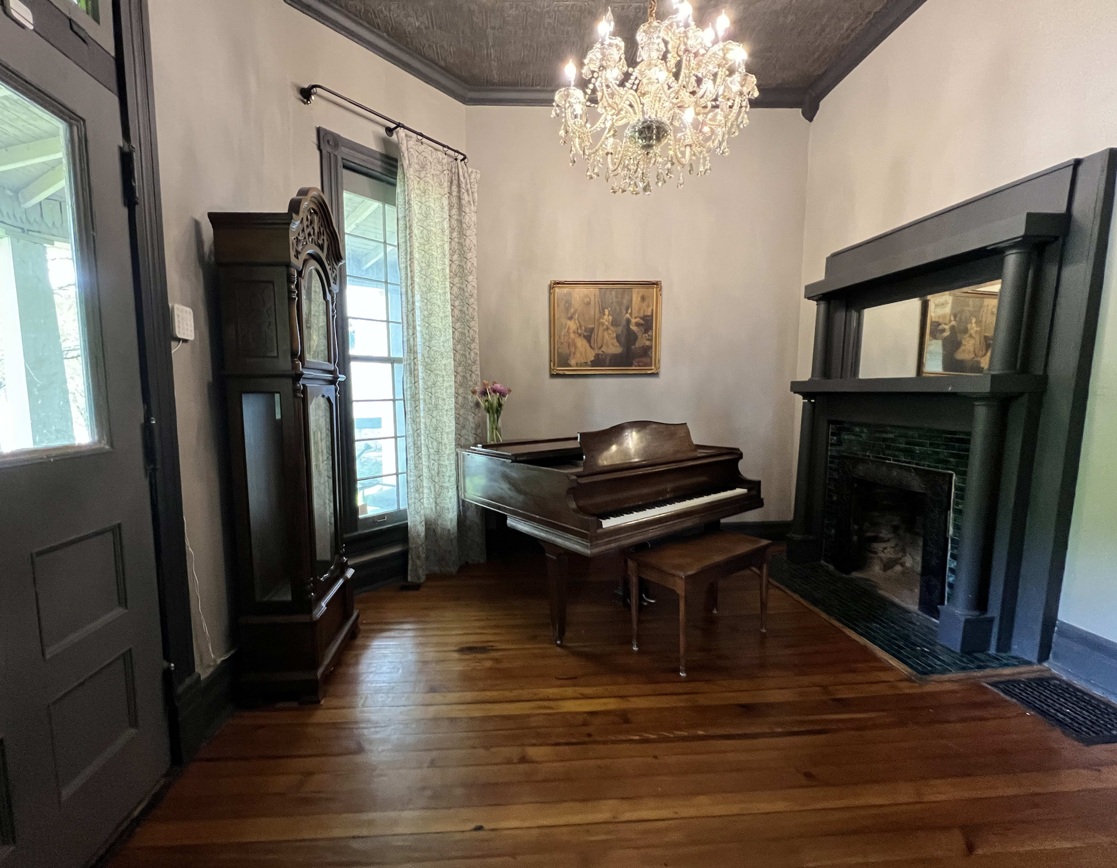 The image shows a room featuring a grand piano, an ornate chandelier, an antique cabinet, and a fireplace, with wooden flooring and a large window.