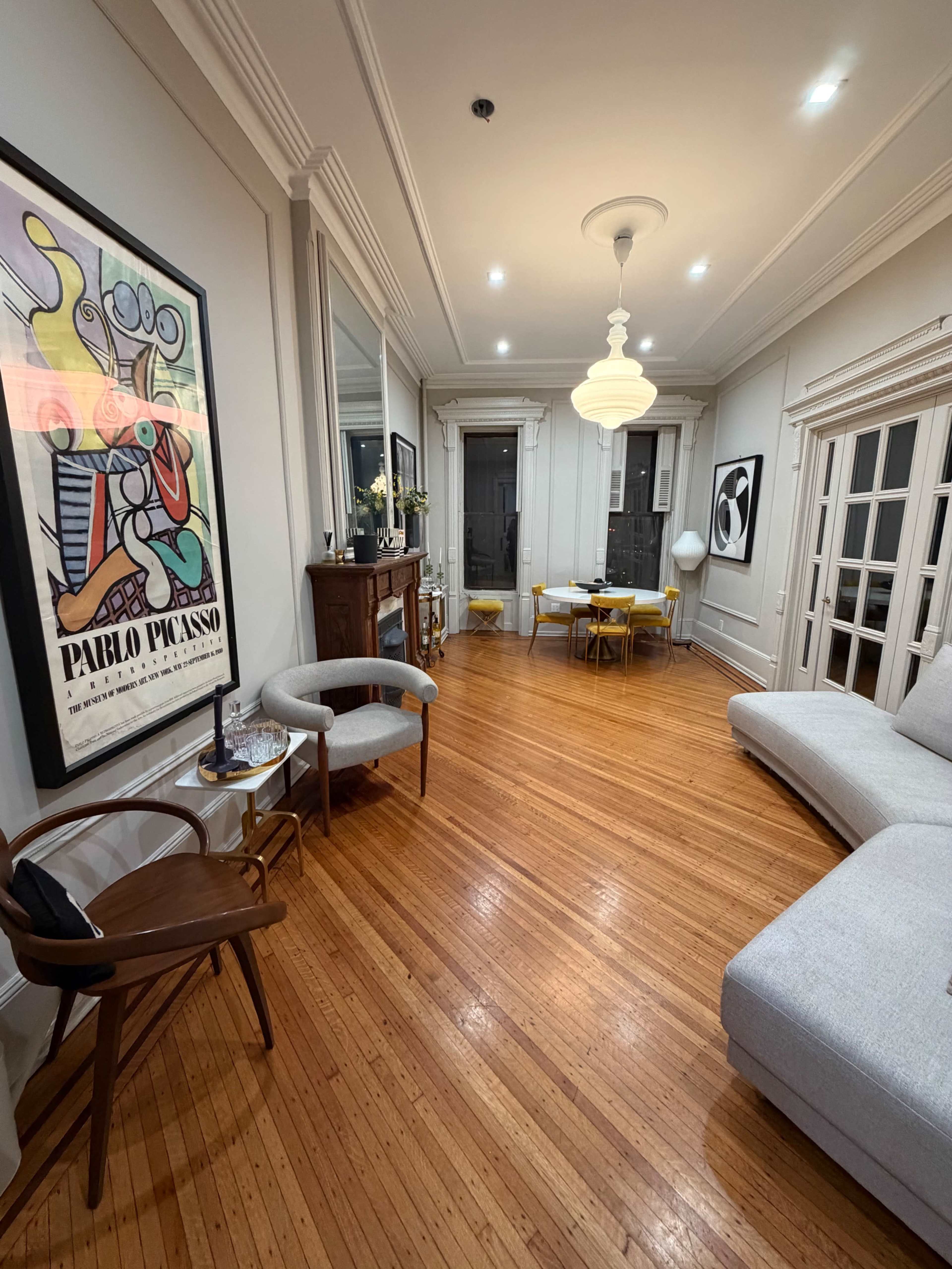 The room features hardwood floors, a gray sectional sofa, a wooden side table, a decorative poster, and a dining area with a round table and yellow chairs.