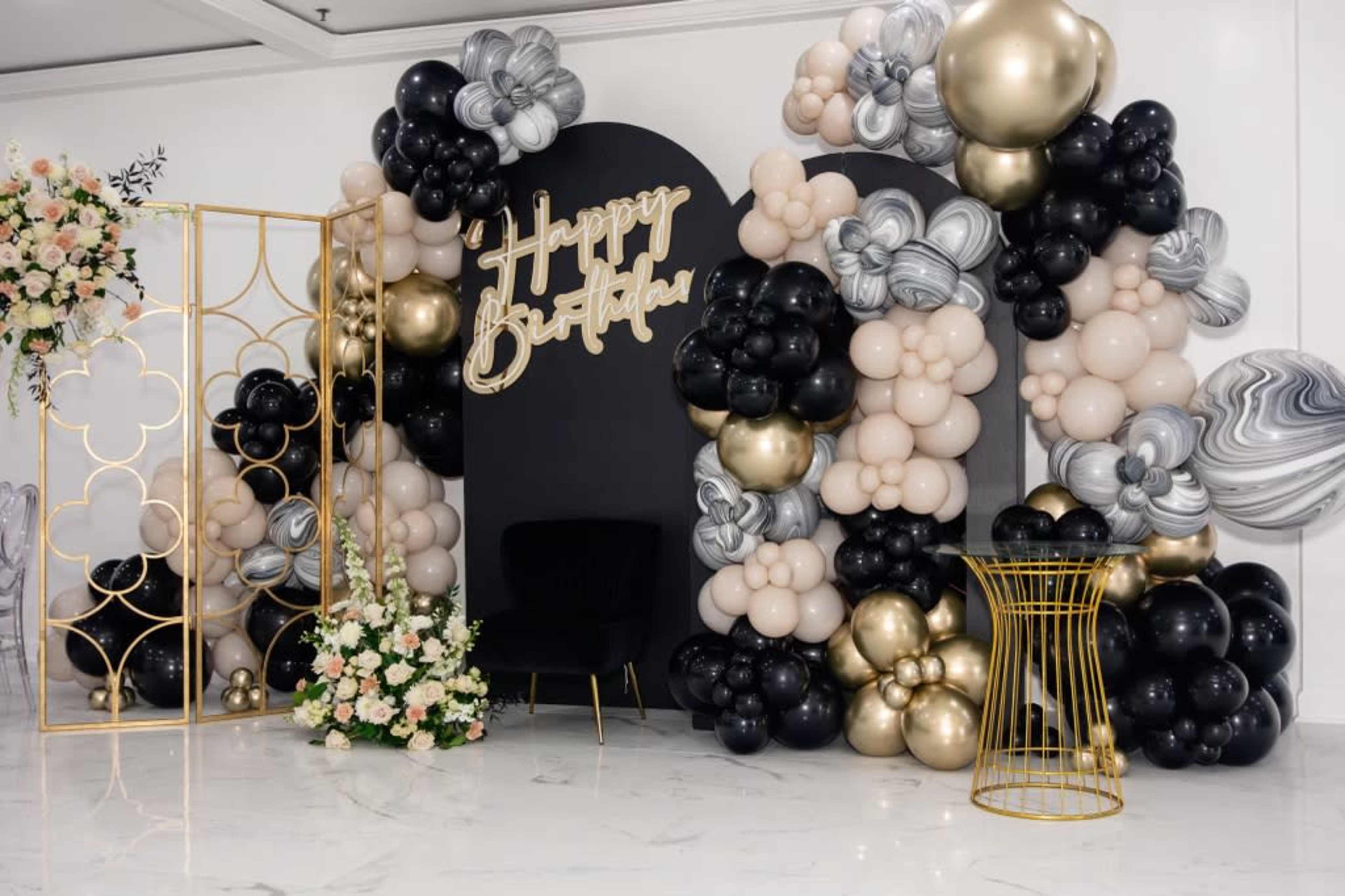The scene features a decorated birthday backdrop with black, gold, and marble-patterned balloons, along with floral arrangements and a black seating area.