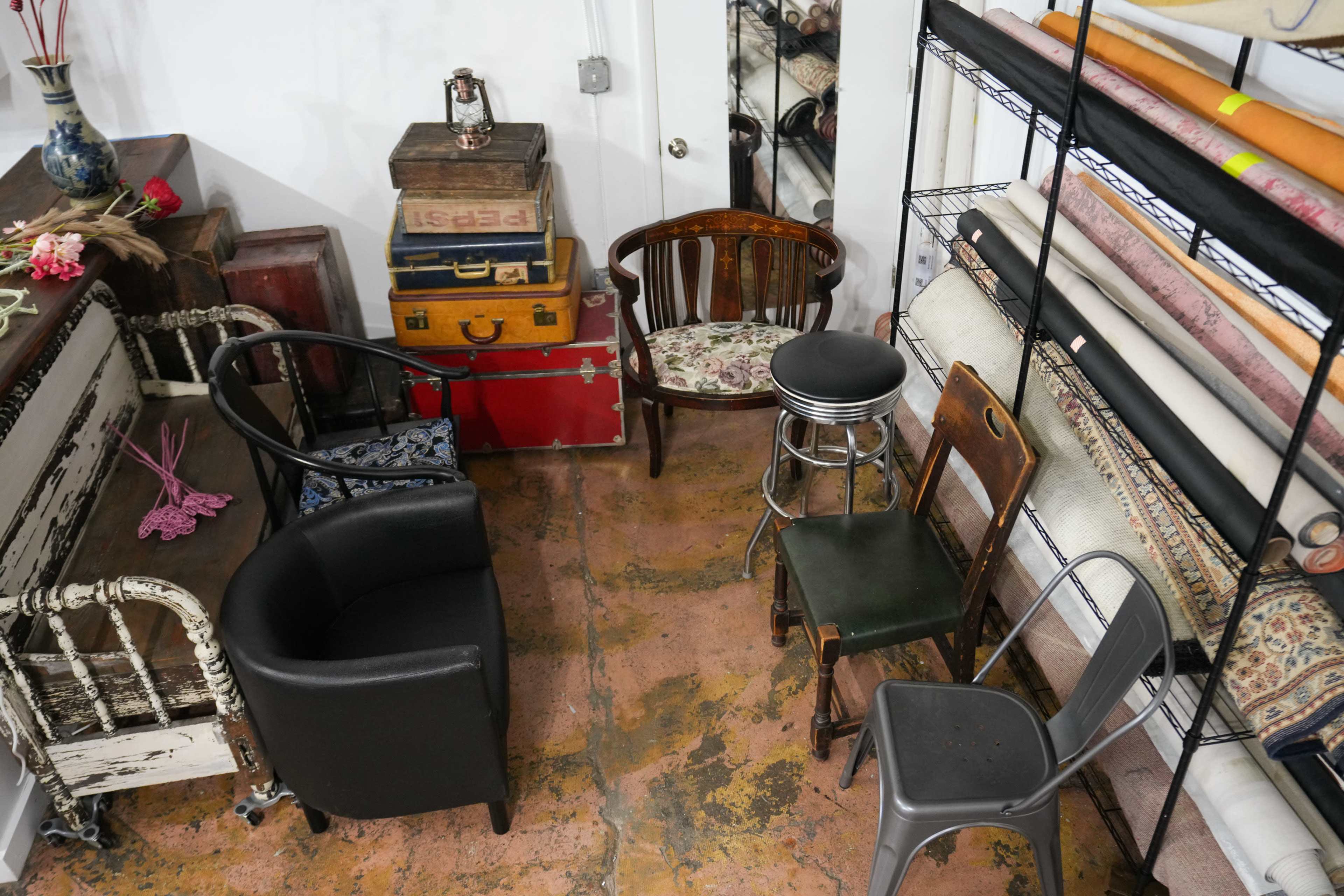 The image shows a small room with various types of chairs arranged around a collection of vintage furniture and rolls of fabric stacked on shelves.