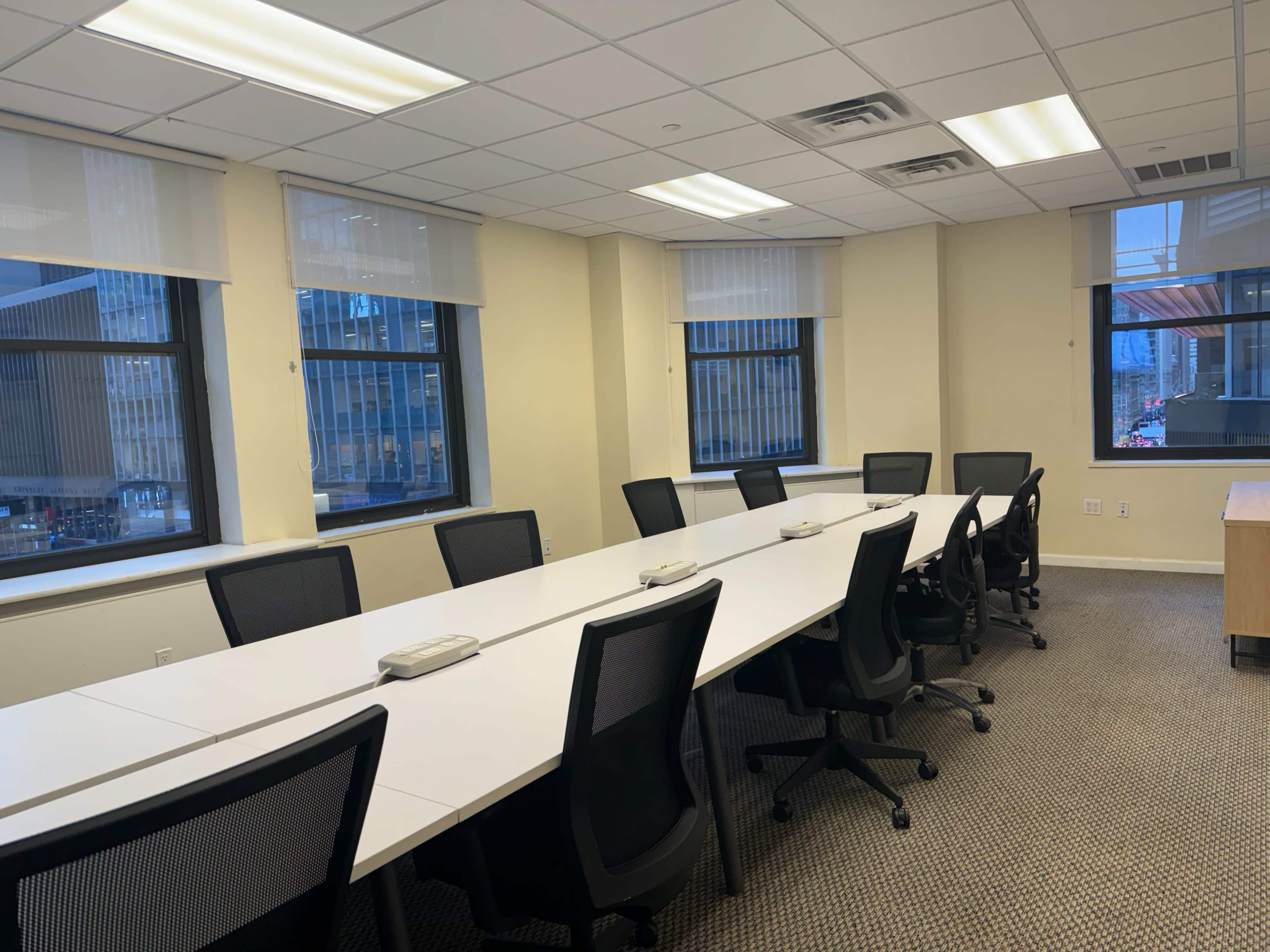 A conference room features a long white table surrounded by black office chairs, with large windows providing views of the city.