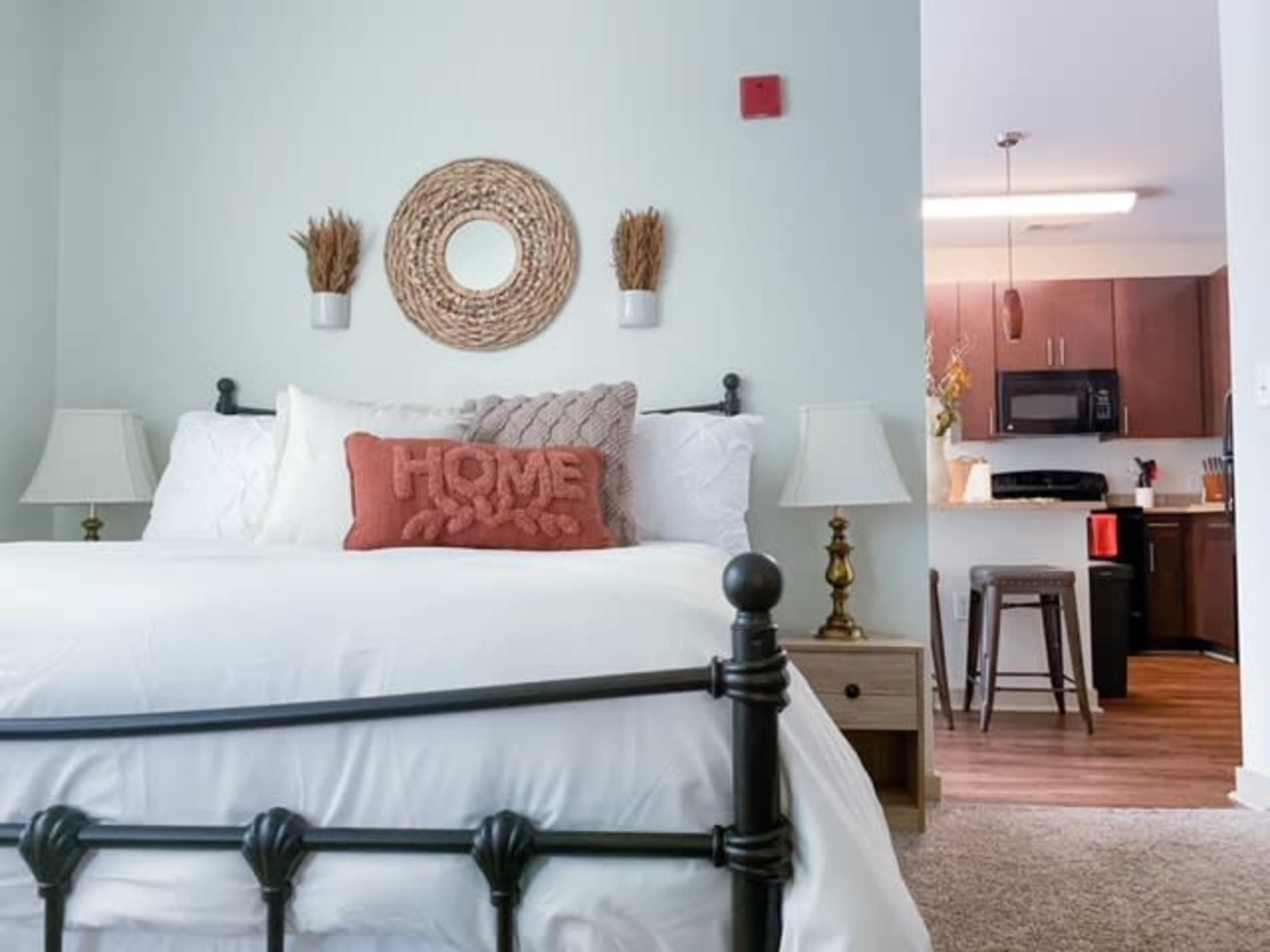 A well-decorated bedroom features a dark metal bed frame with white bedding, adorned with a decorative pillow, and leads to a kitchen area in the background.