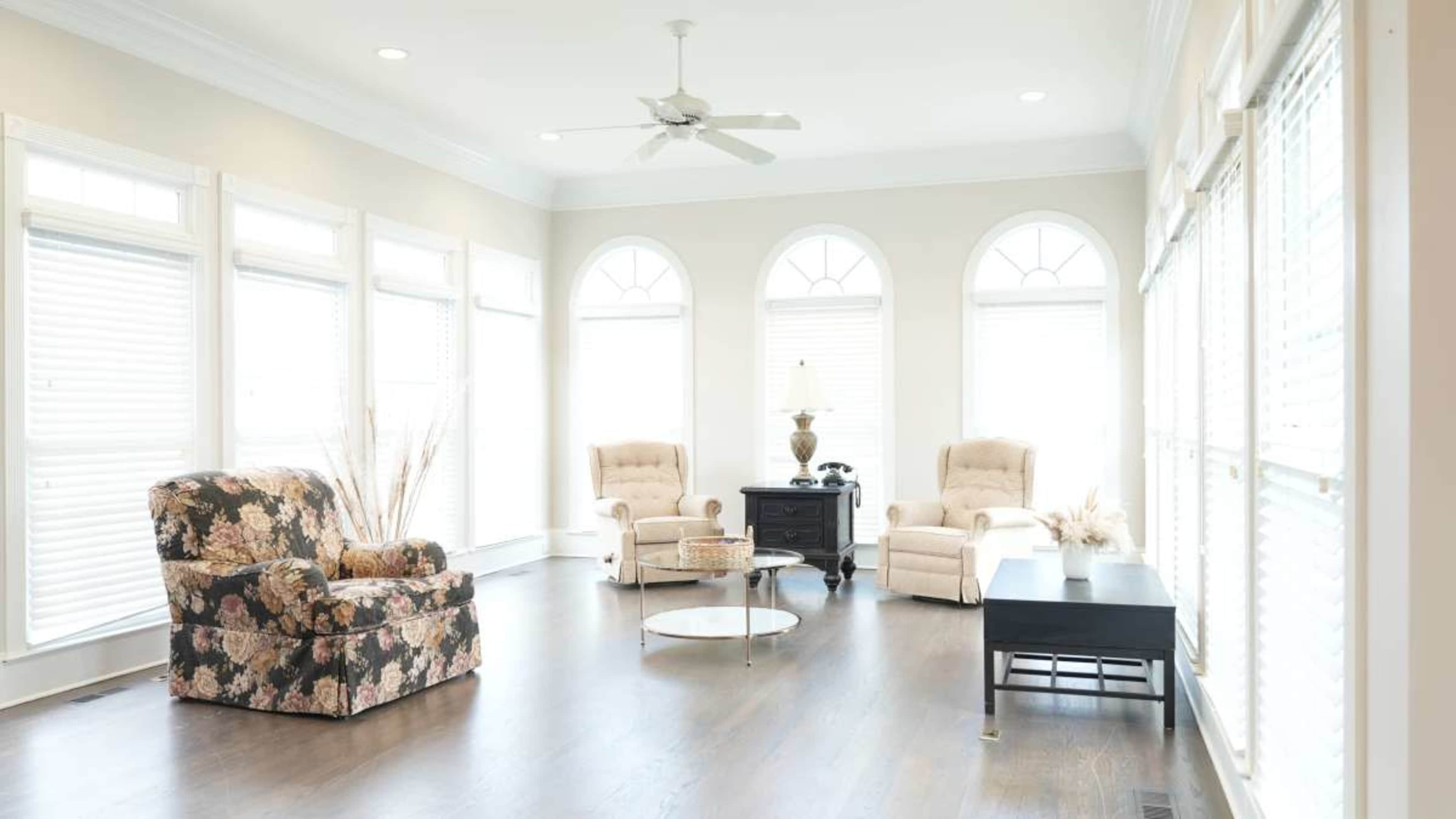 A bright, airy living room features large windows with arched tops, two beige recliners, a floral-patterned armchair, a round coffee table, and a black side table.