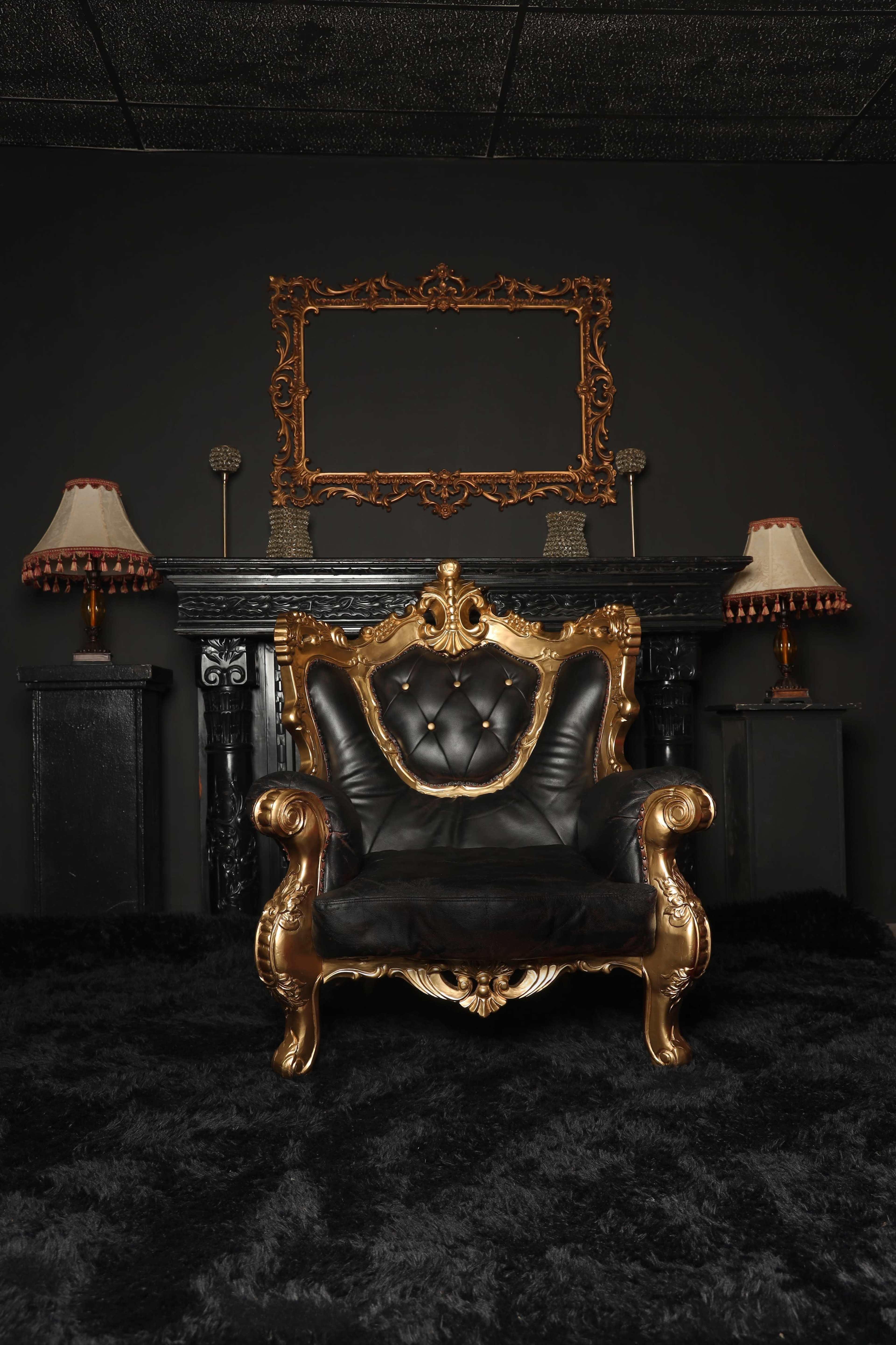 The image shows a plush, black armchair with ornate gold detailing positioned against a black wall and a decorative fireplace, accompanied by two elegant lamps and a gilded mirror.