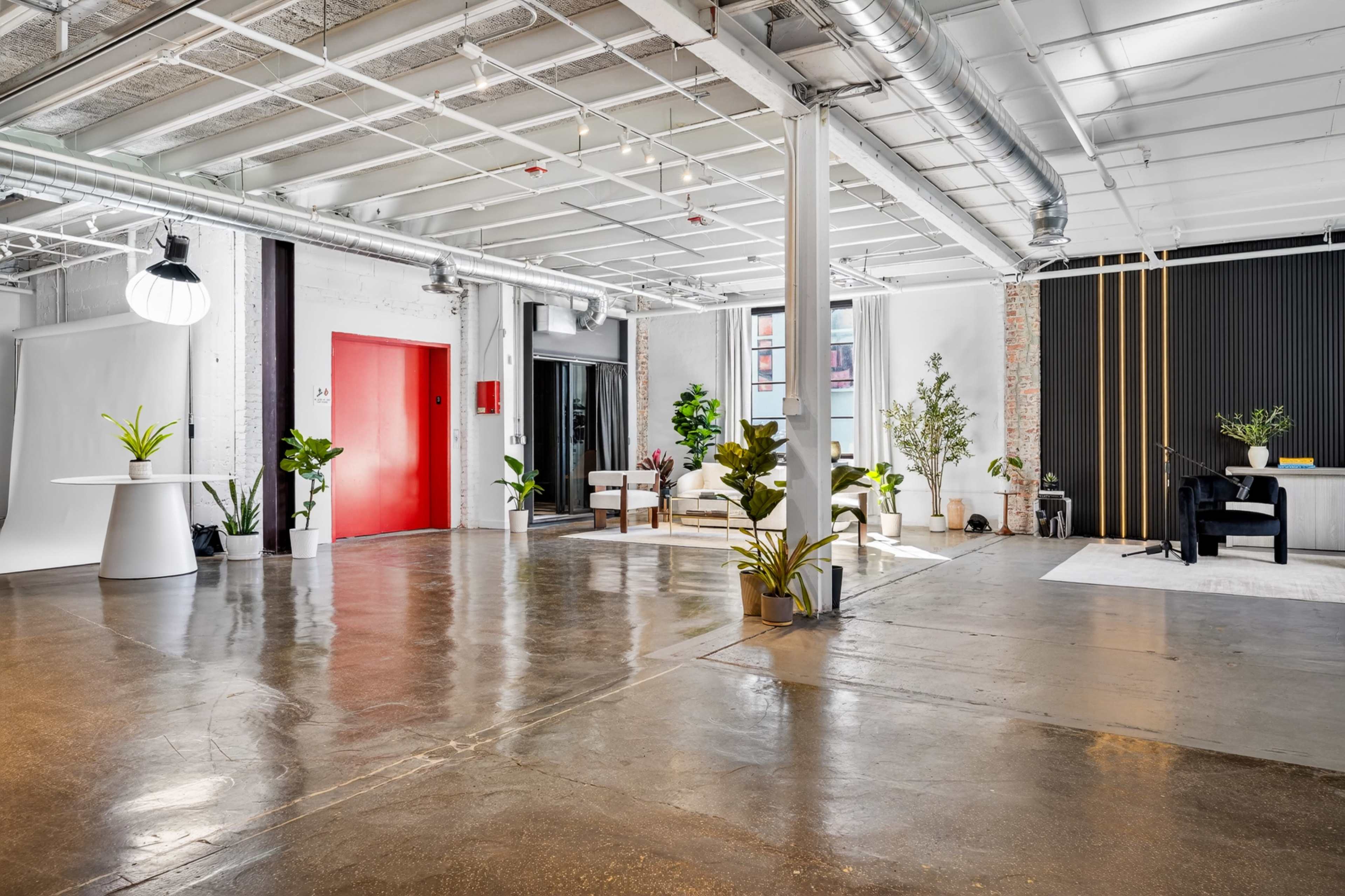 The image shows a bright, spacious interior featuring polished concrete floors, white walls, plants, and distinct areas for seating and work, with a red door and large windows providing natural light.