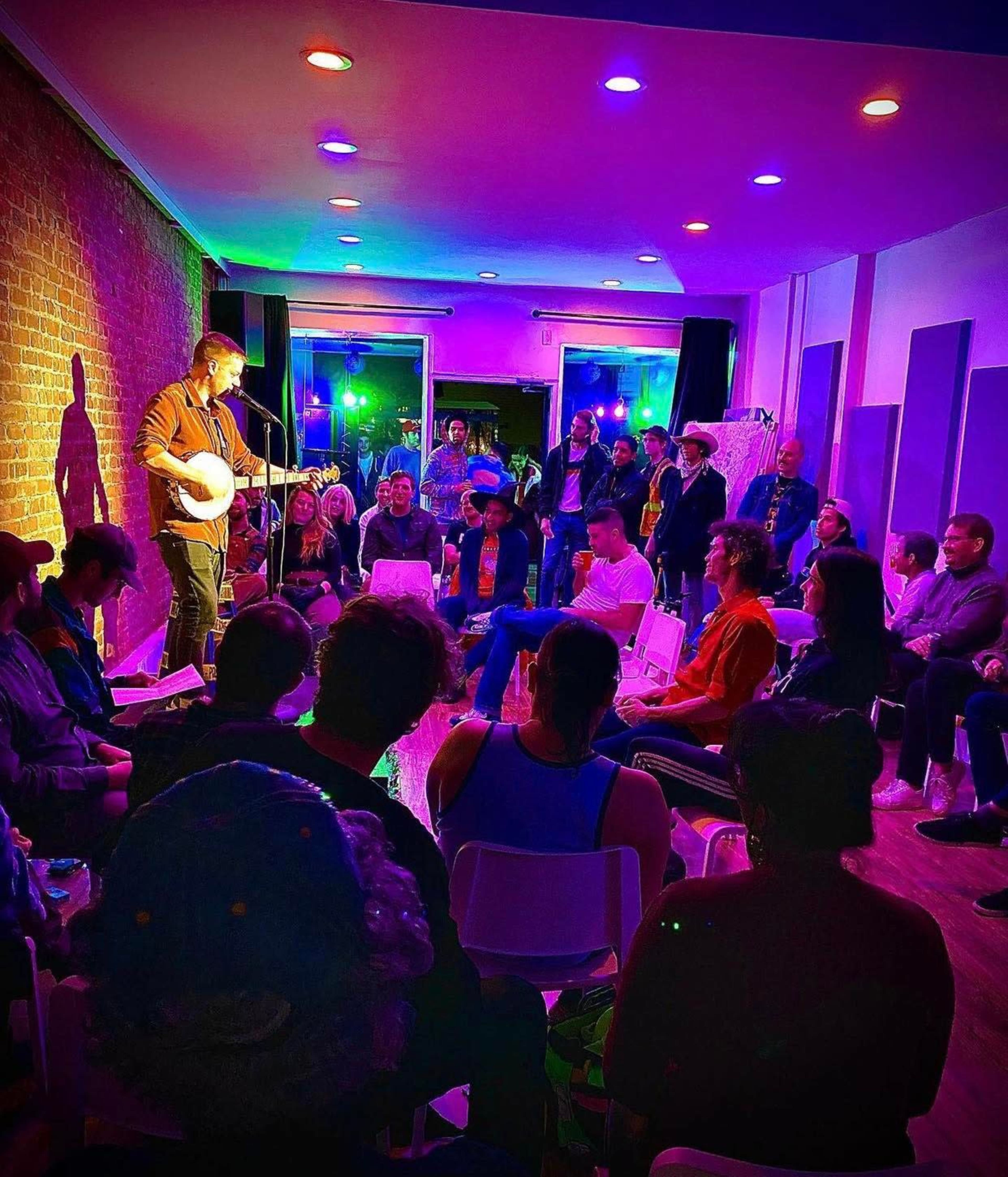 A musician performs with a banjo in front of an audience seated in a brightly lit room with colorful stage lighting.