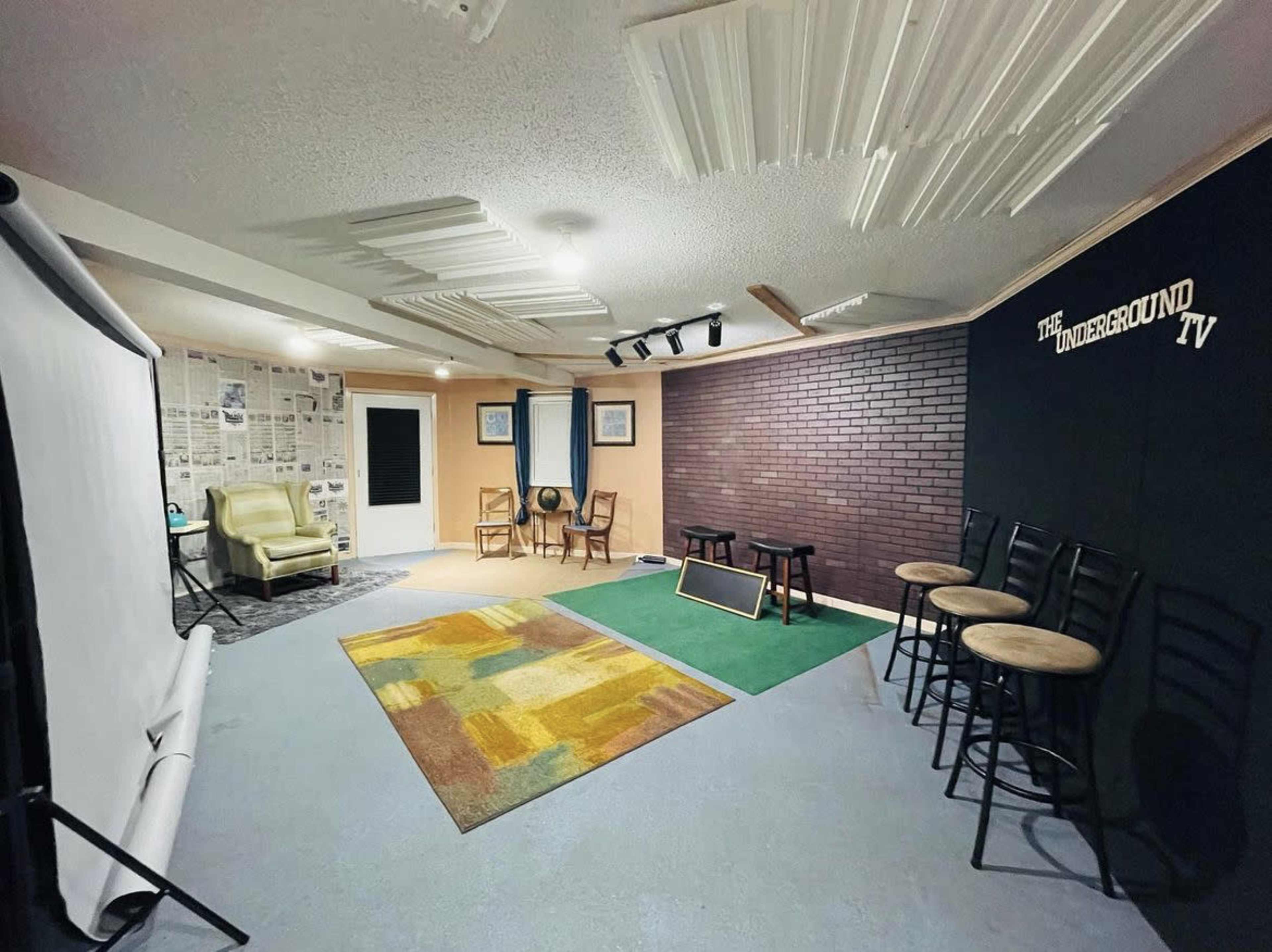 A studio space featuring a carpet, a green area, seating arrangements, a white backdrop, and walls decorated with bricks and newsprint.