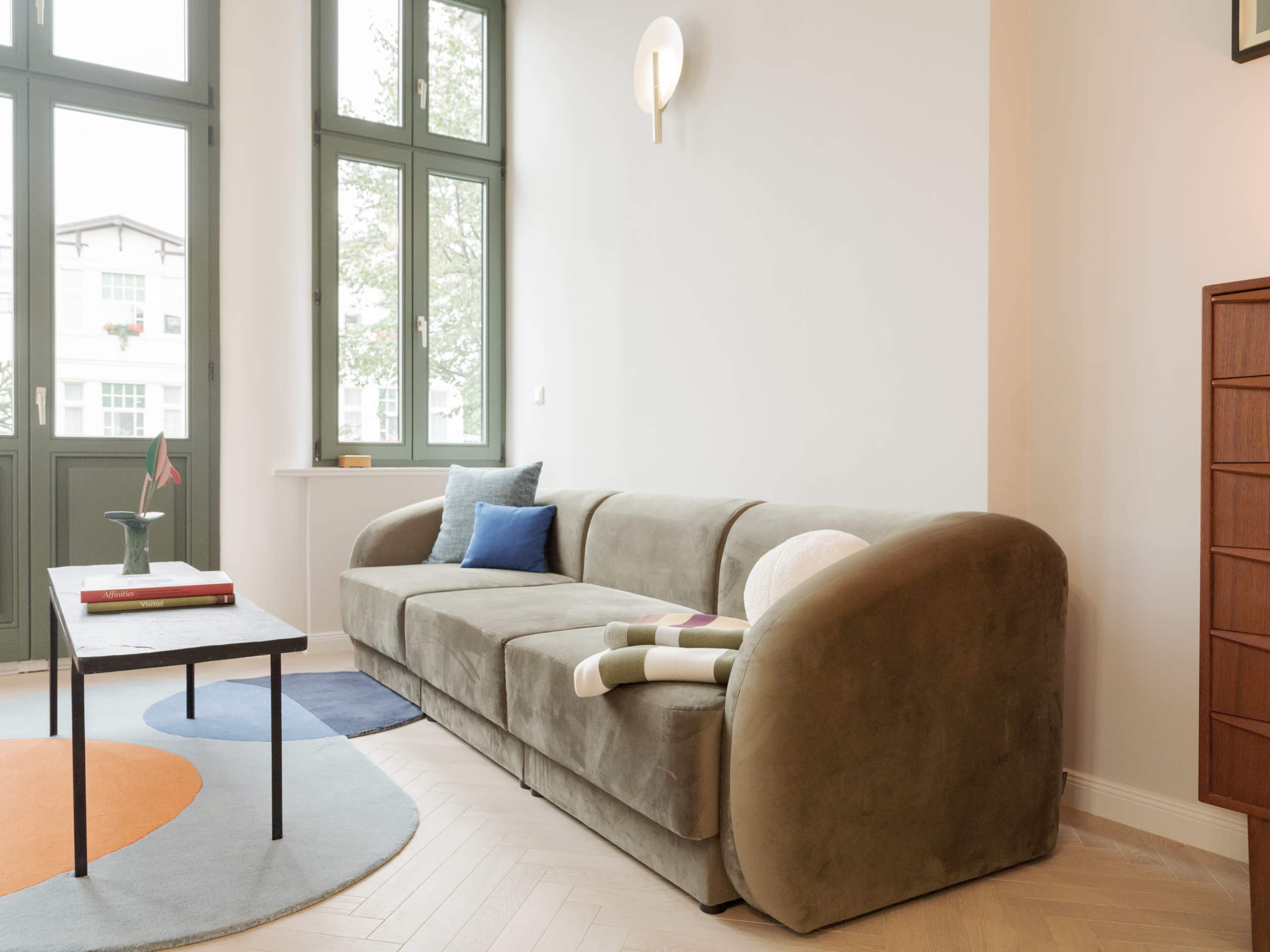 A green couch with rounded edges is positioned near a coffee table in a well-lit room featuring large windows.