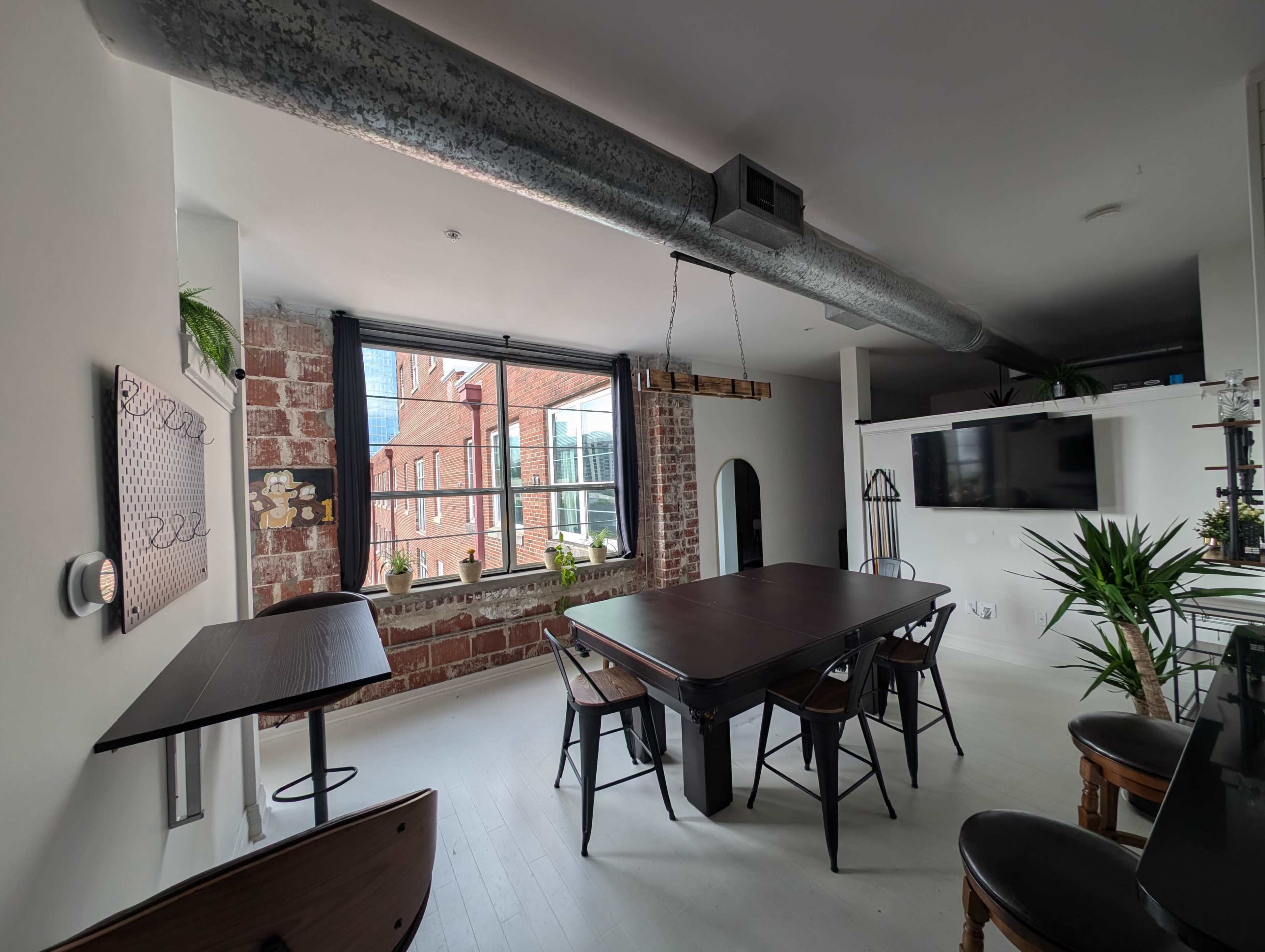 The image shows a modern dining area with a dark wooden table and black chairs, exposed brick walls, and large windows letting in natural light.