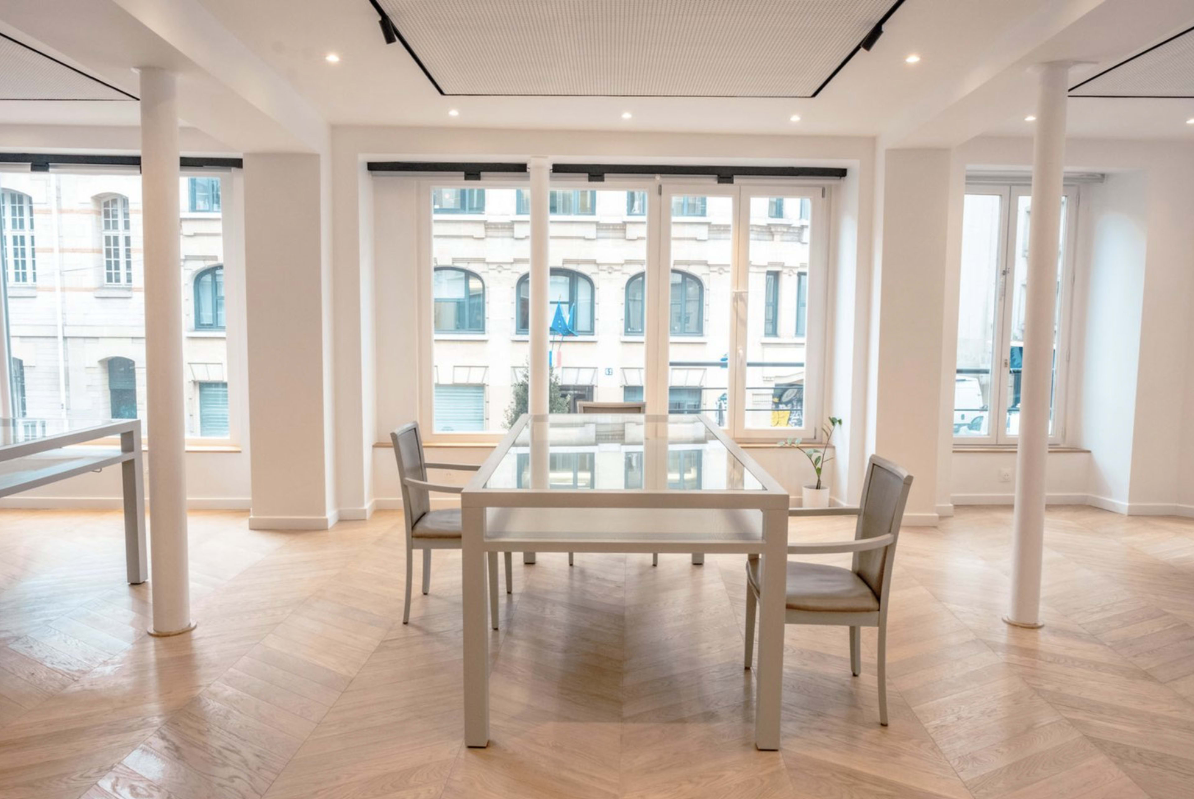 A bright, modern conference room features a glass table surrounded by four chairs, with large windows showcasing a view of a building across the street.