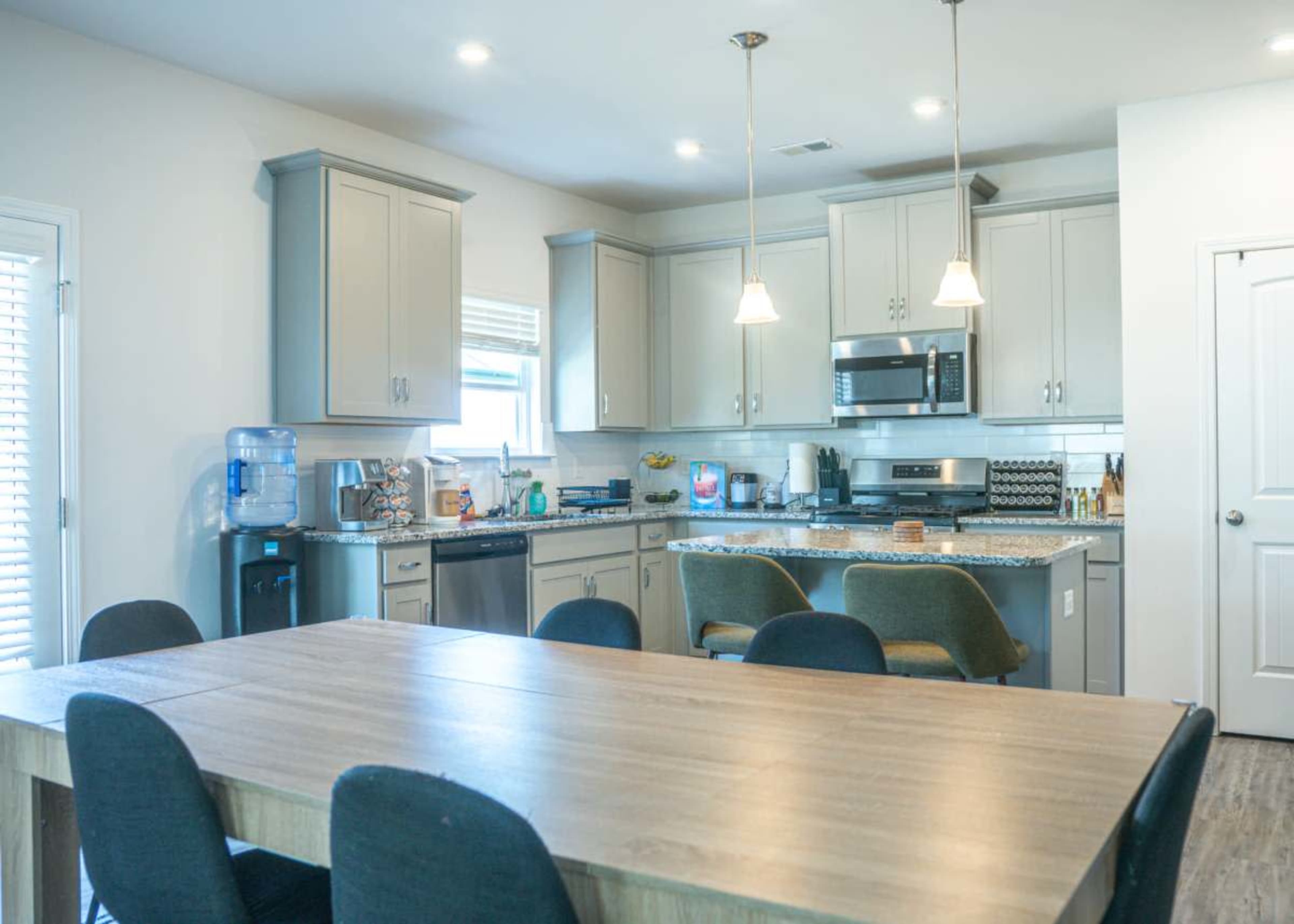 A modern kitchen with gray cabinets and granite countertops features a dining table with black chairs and an array of kitchen appliances.