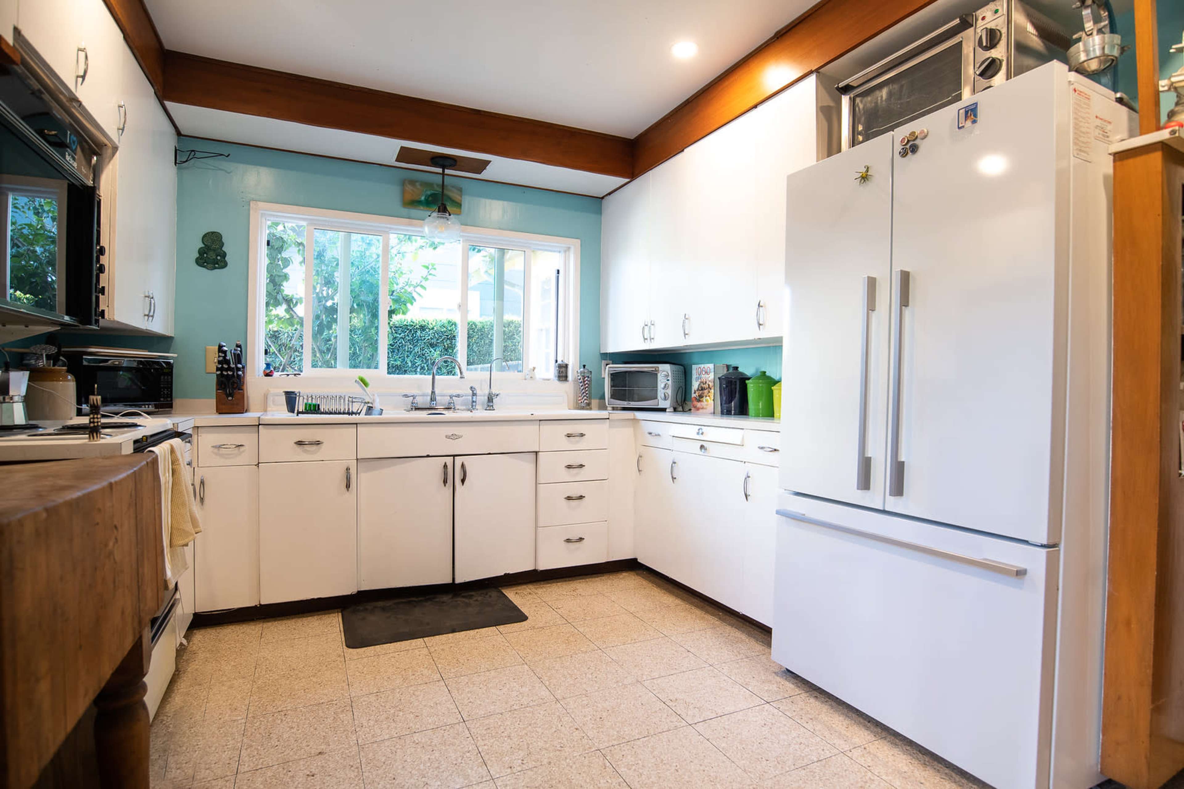 The image shows a bright kitchen with white cabinets, a large refrigerator, and a window overlooking an outdoor area.