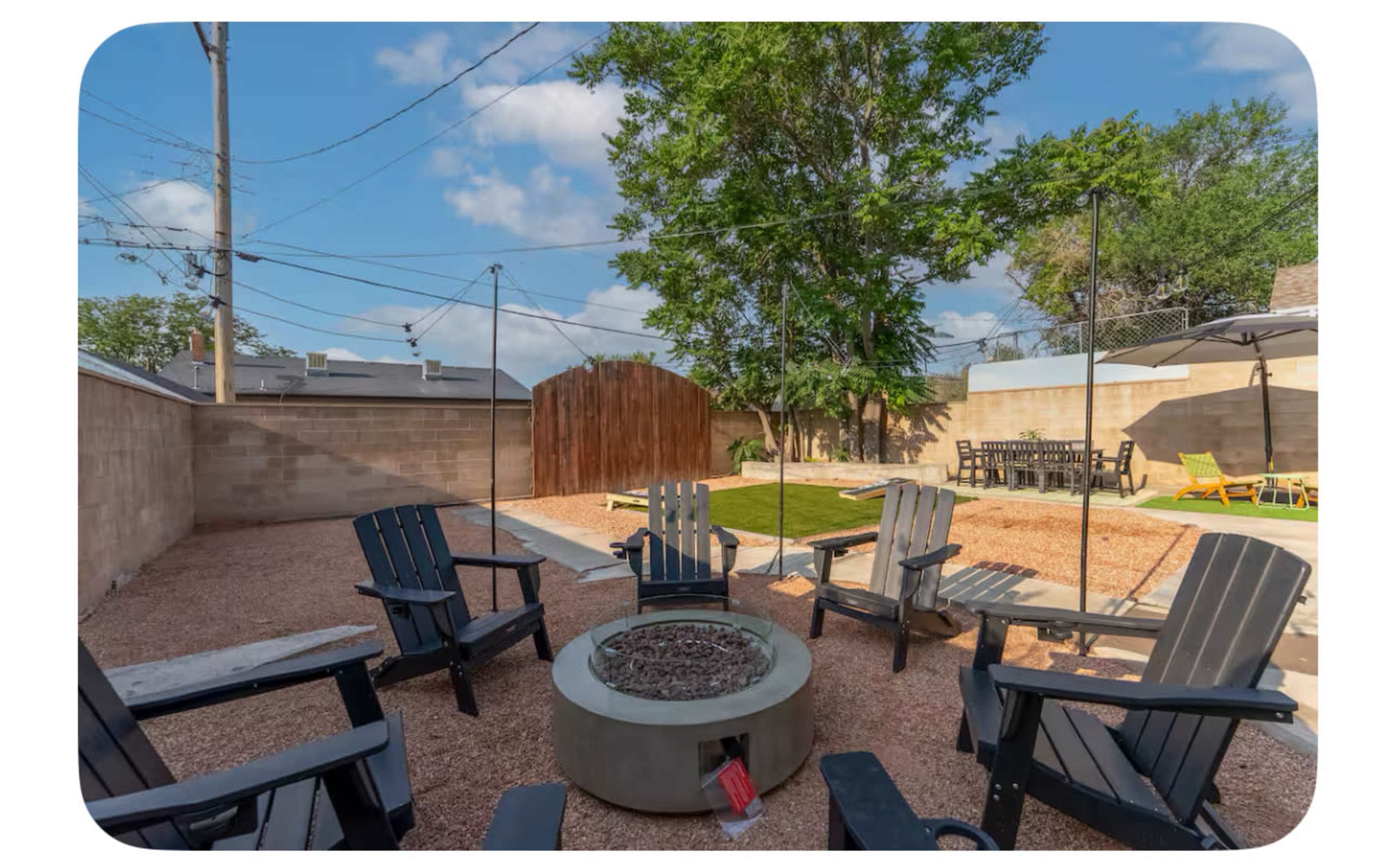 The image shows a backyard with a fire pit surrounded by black outdoor chairs, a grassy area, and a wooden fence in the background.
