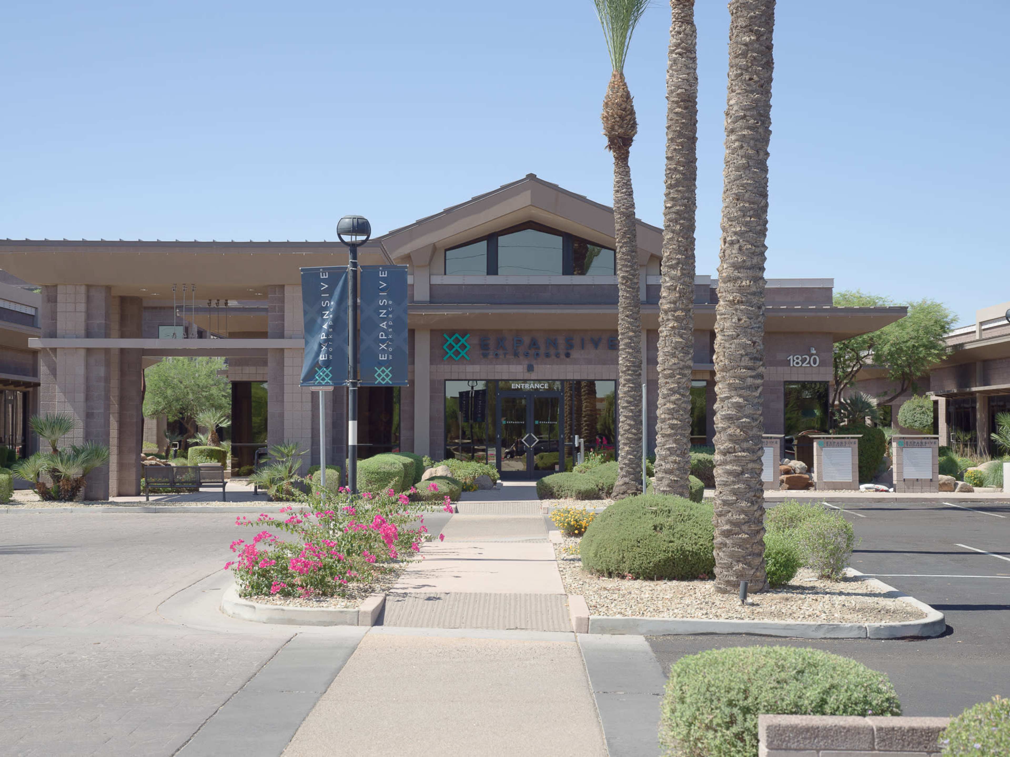 The image depicts a business building with a modern facade surrounded by palm trees and landscaped gardens, located on a paved pathway.