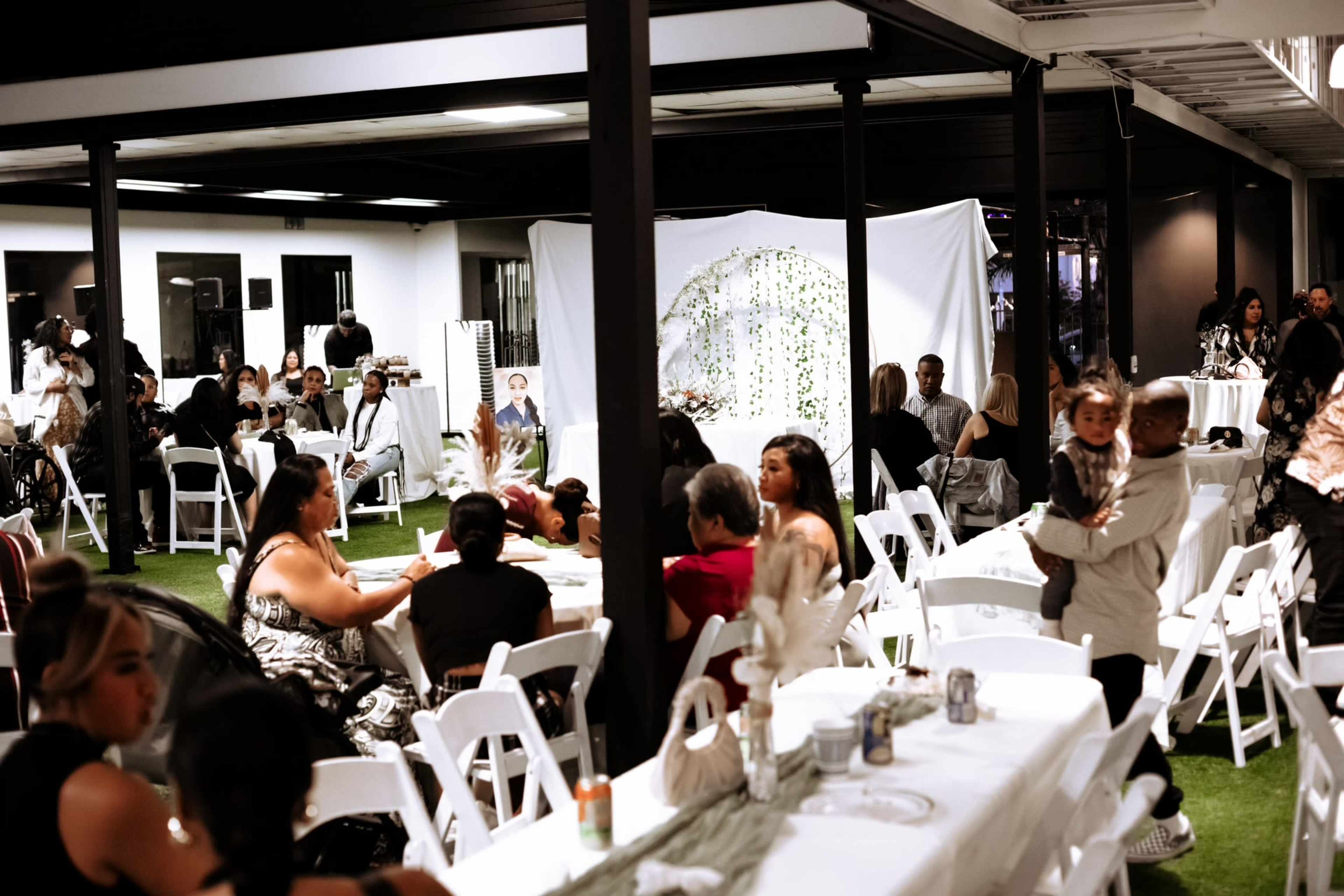 The image shows a lively indoor event space with tables arranged for dining and guests engaged in conversation, surrounded by decorations and a backdrop featuring greenery.