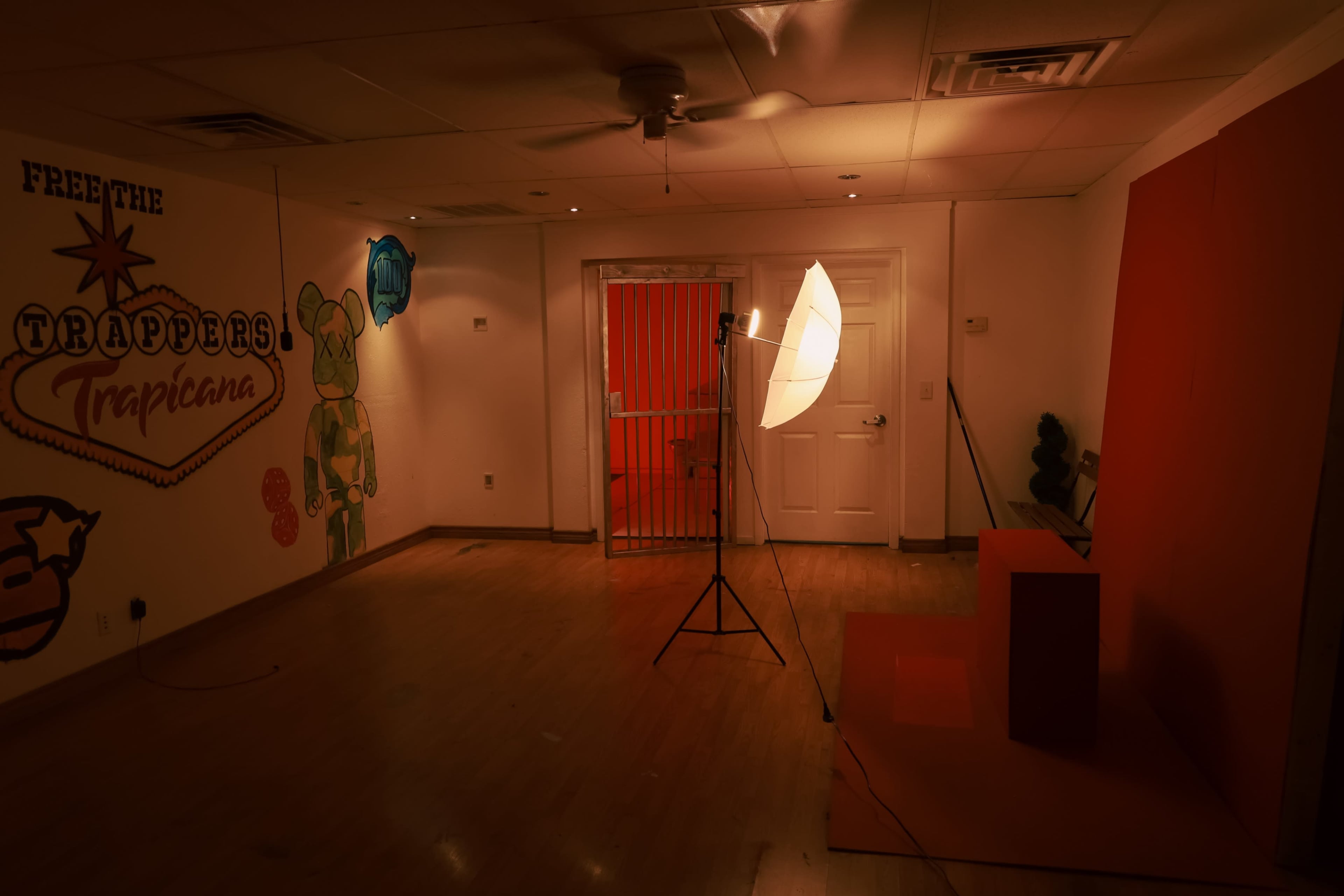 The image depicts an empty room with a backdrop and lighting setup, featuring a graffiti mural on one wall and a barred door on the opposite side.