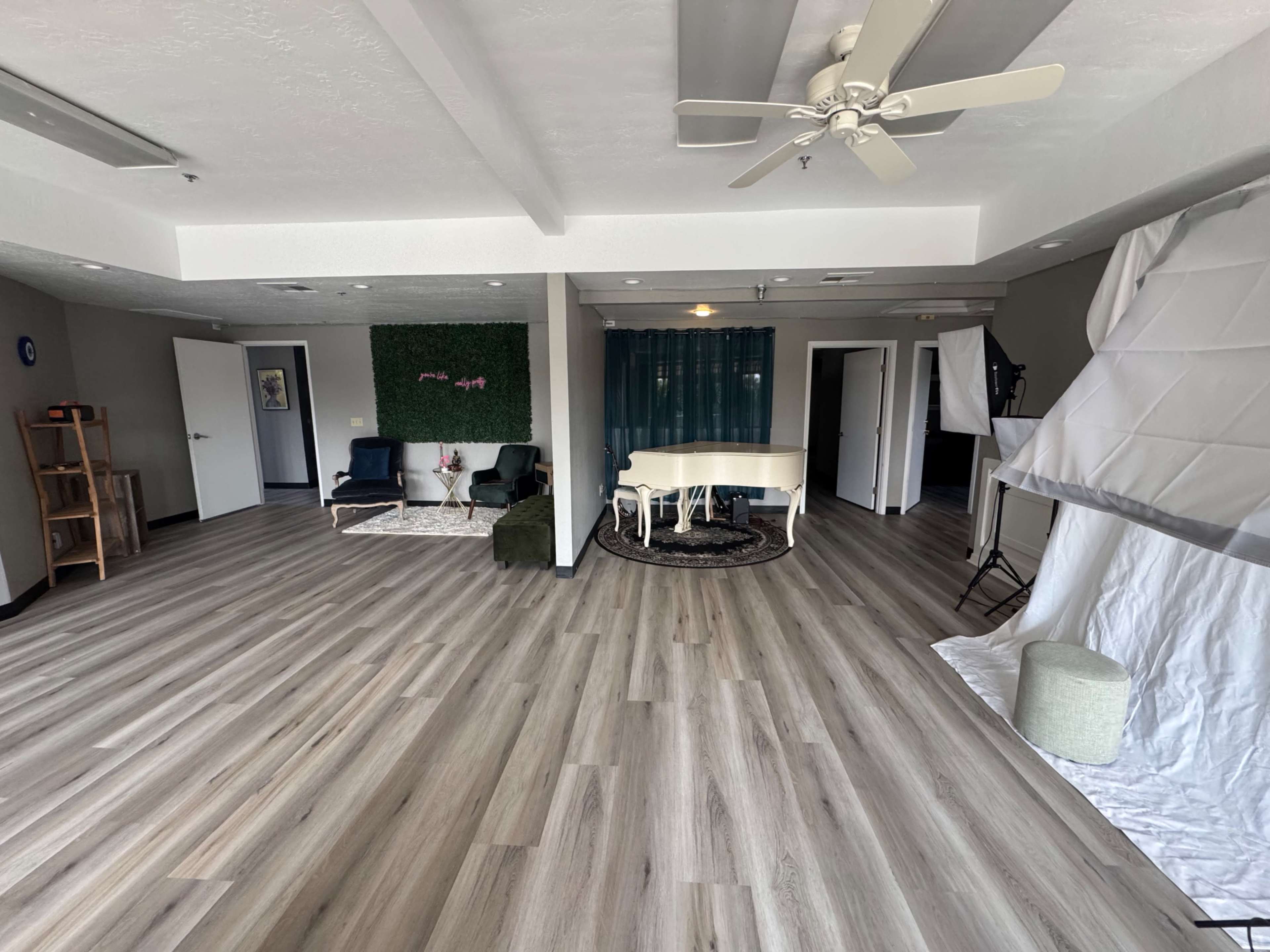 The image shows a spacious interior with wooden flooring, a piano in the center, and various seating areas along with green decor and studio elements.