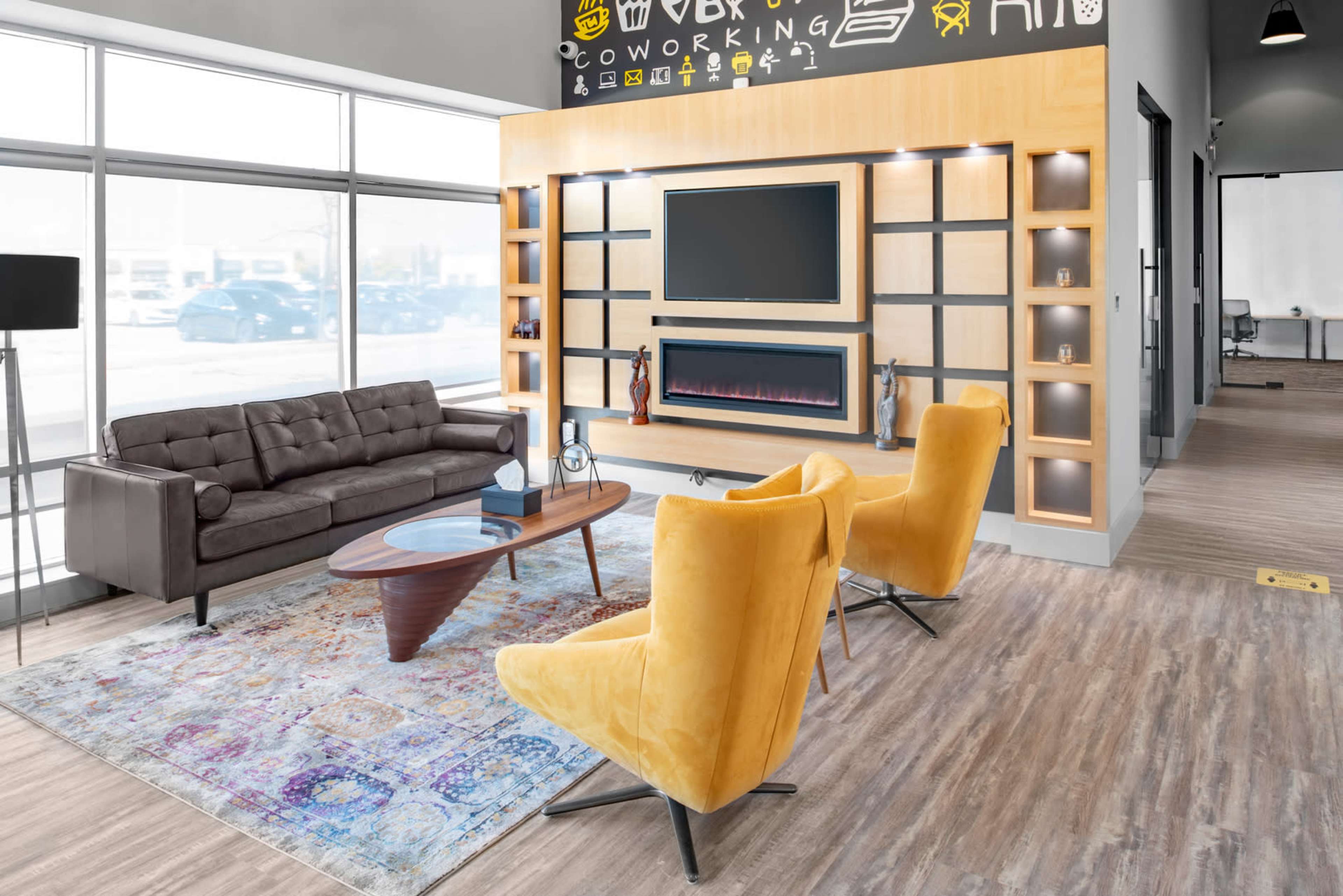 A modern coworking space featuring a brown sofa, two yellow armchairs, a coffee table, and a wall-mounted TV above a decorative shelf.
