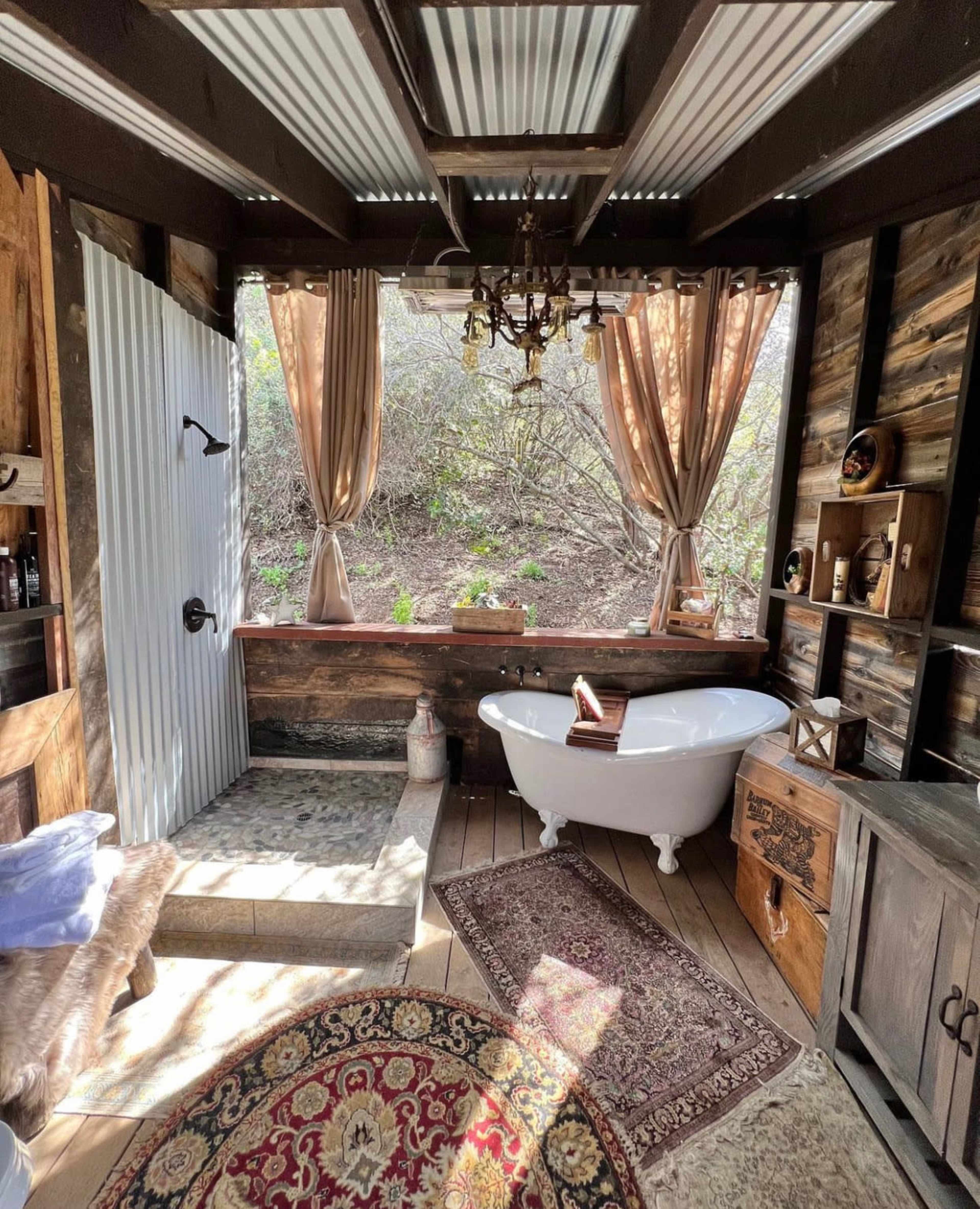 A rustic bathroom featuring a freestanding bathtub, a vintage chandelier, wooden walls, and large windows framed by curtains.