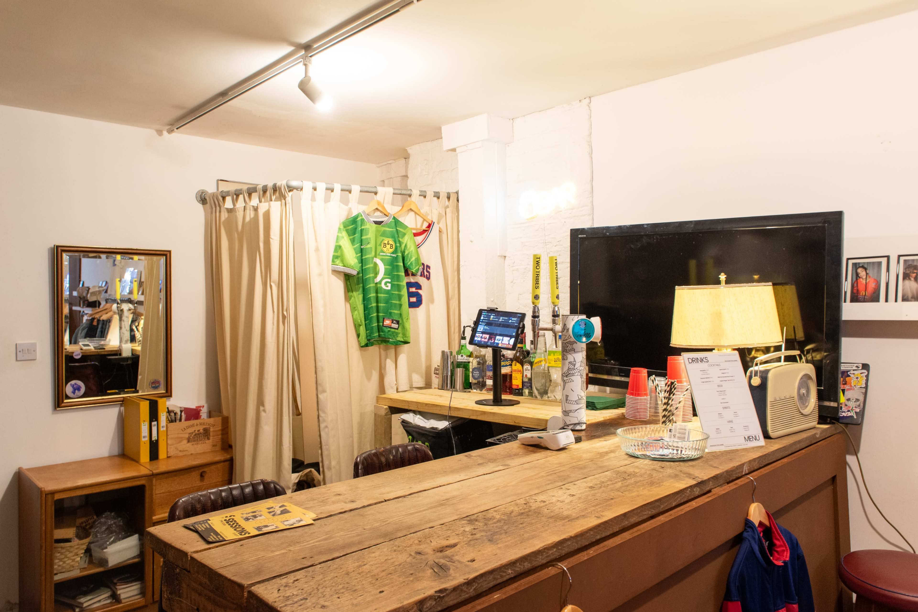 The image shows a small, cozy bar area with a wooden counter, a dimly lit lamp, and various items displayed on shelves, including a sports jersey hanging on the wall.
