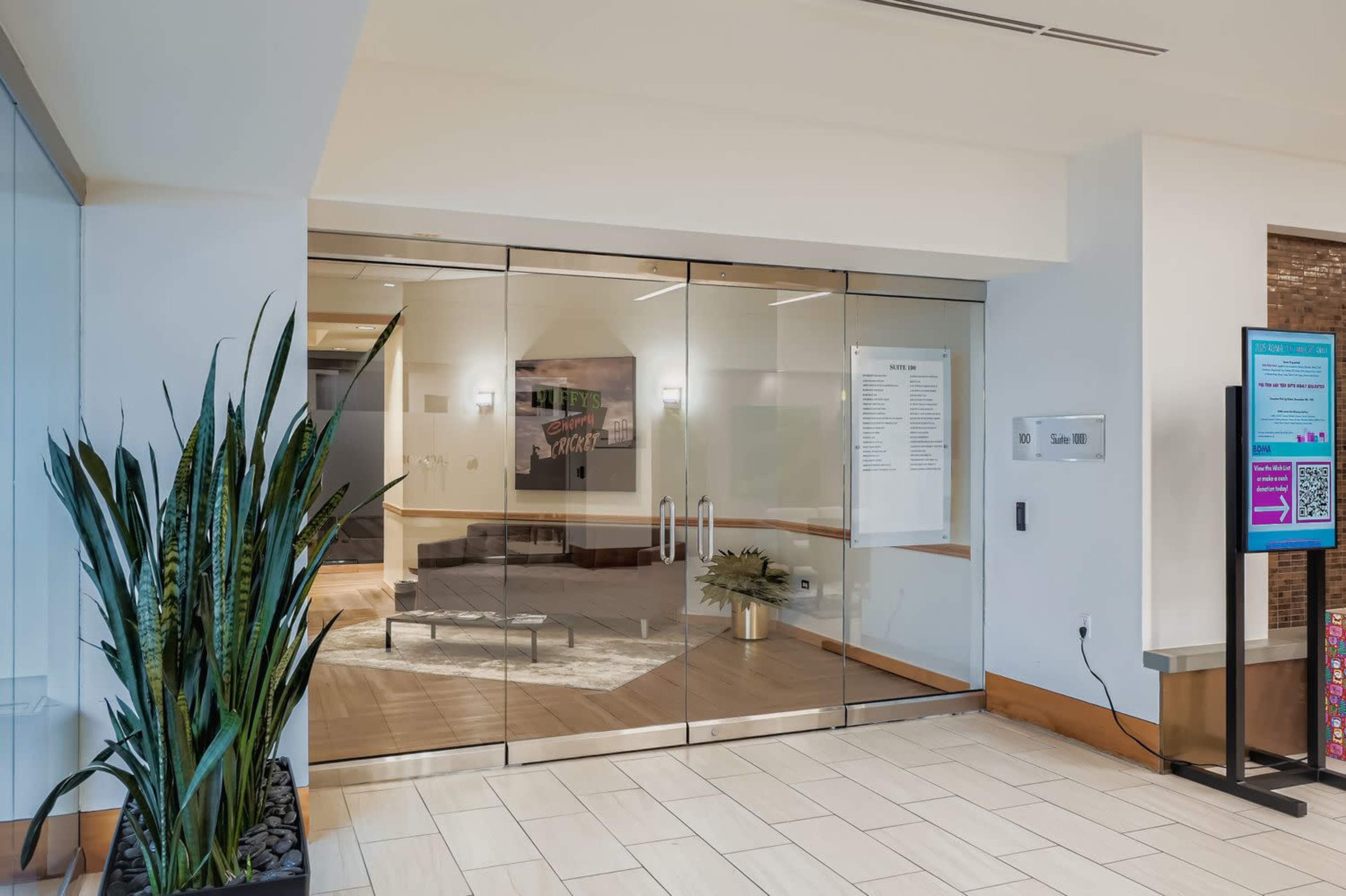 The image shows a modern entrance with glass doors leading to a room, accompanied by a plant and digital signage to the side.
