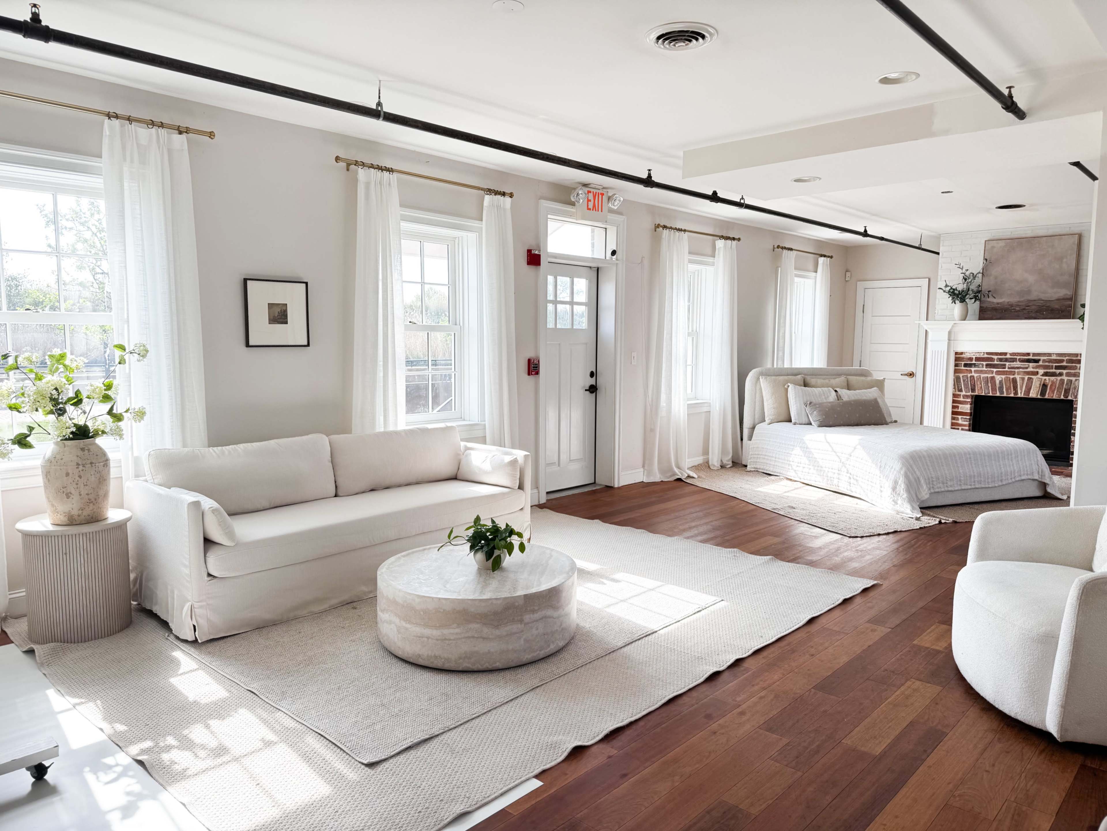 The image shows a bright, modern interior space featuring a living area with a white sofa, a round coffee table, and a bed in a cozy corner, accented by large windows and light curtains.