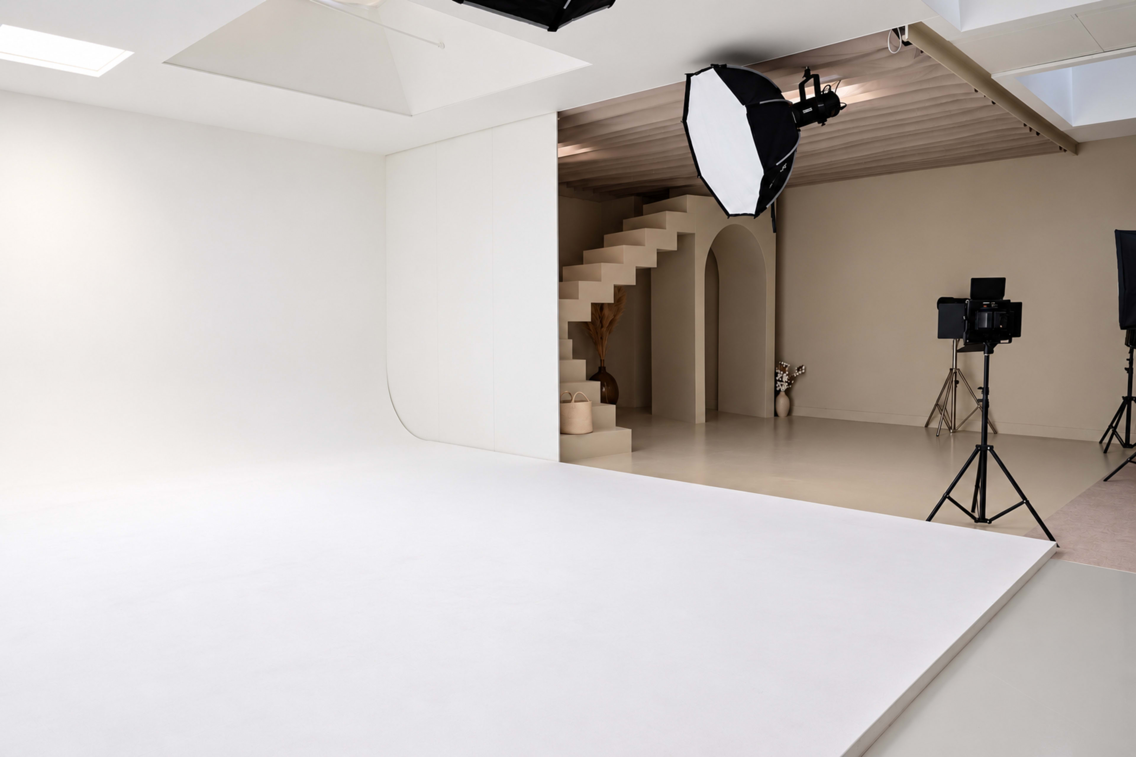 The image shows a minimalist photo studio with a white backdrop, a spiral staircase in the background, and professional lighting equipment set up.