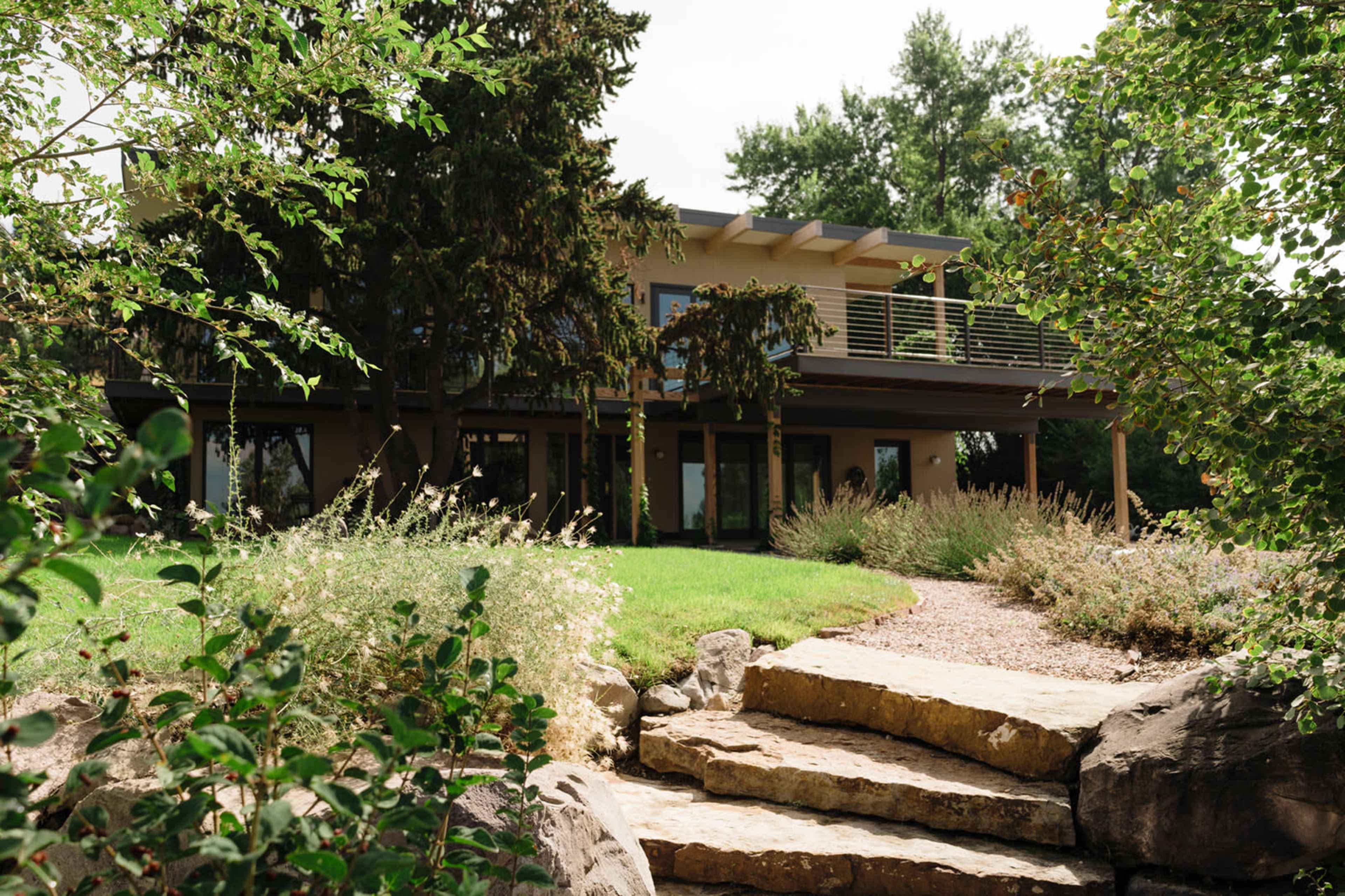 A modern house with a balcony is surrounded by greenery and rocks in a landscaped yard.