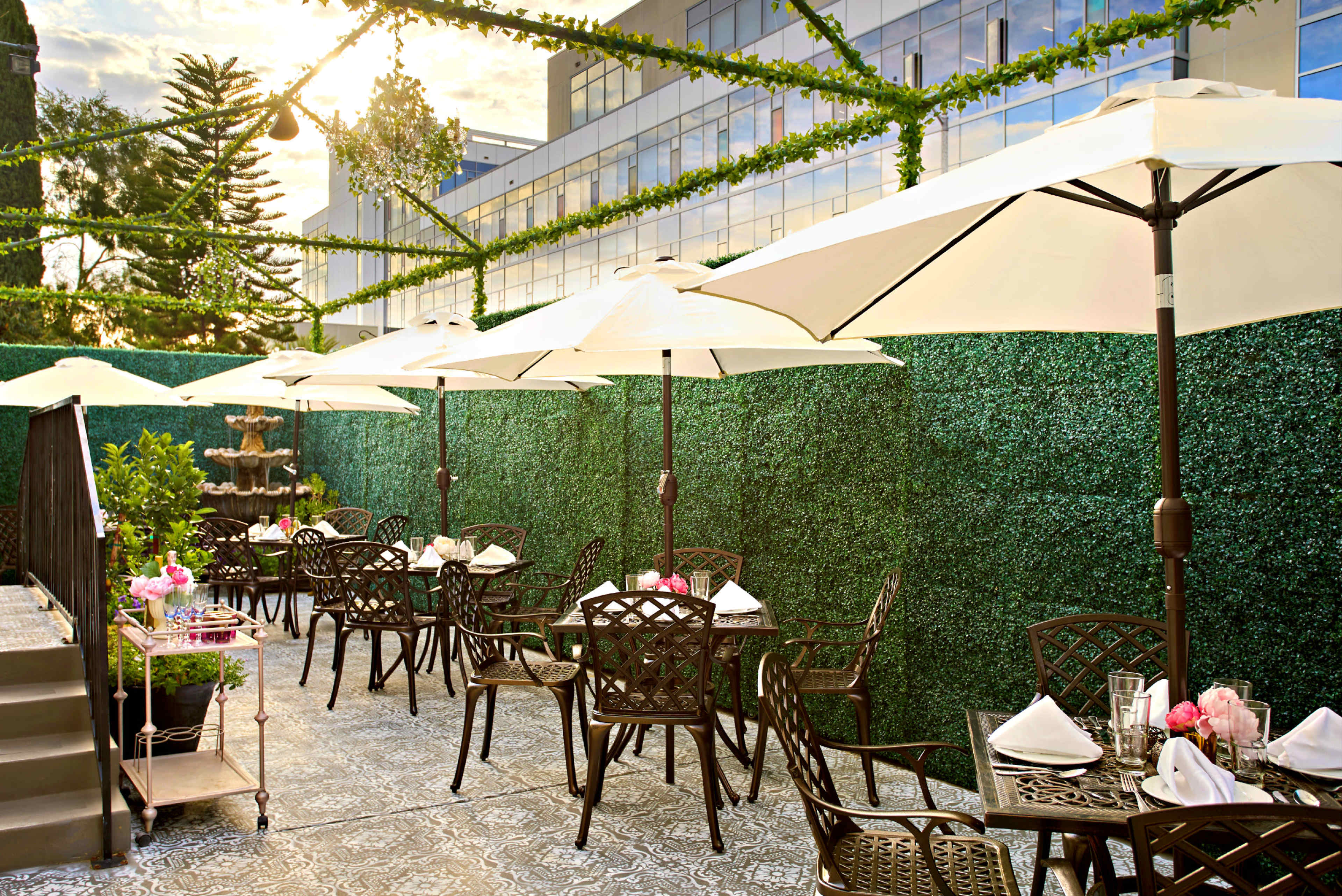 An outdoor dining area with tables set under large umbrellas, surrounded by lush green walls and a fountain in the background.